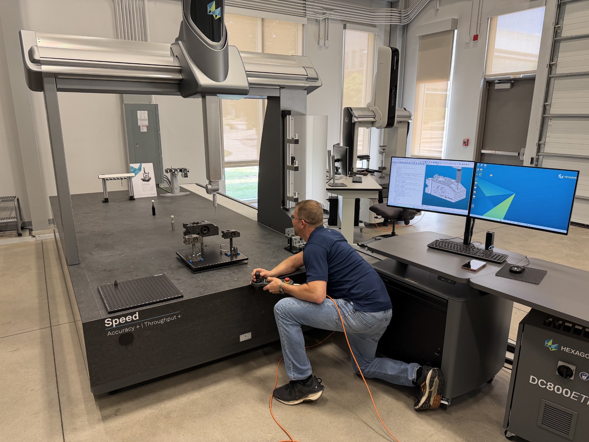 The photo shows a large, high‑precision industrial measurement machine inside a clean, modern lab or manufacturing facility. The machine has a wide granite base labeled “Speed – Accuracy + Throughput +” and is equipped with a tall, overhead gantry system that holds a coordinate‑measuring probe positioned above a metal part fixed to the table. Several other small fixtures and tools are arranged on the surface. A person is kneeling beside the machine, holding a controller connected by an orange cable, appearing to operate or calibrate the equipment. To the right, there is a workstation with two computer monitors displaying technical drawings, measurement data, and a blue desktop screen. The workstation includes a keyboard, mouse, and a phone resting on the desk. In the background, there are additional measurement machines, computers, and lab equipment. Large windows and overhead lighting provide bright, even illumination throughout the room. The overall setting appears to be a metrology lab focused on precision inspection or engineering measurement.
