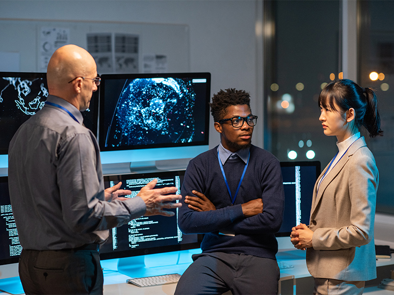 Photo of three people in business formal attire talking in front of monitors
