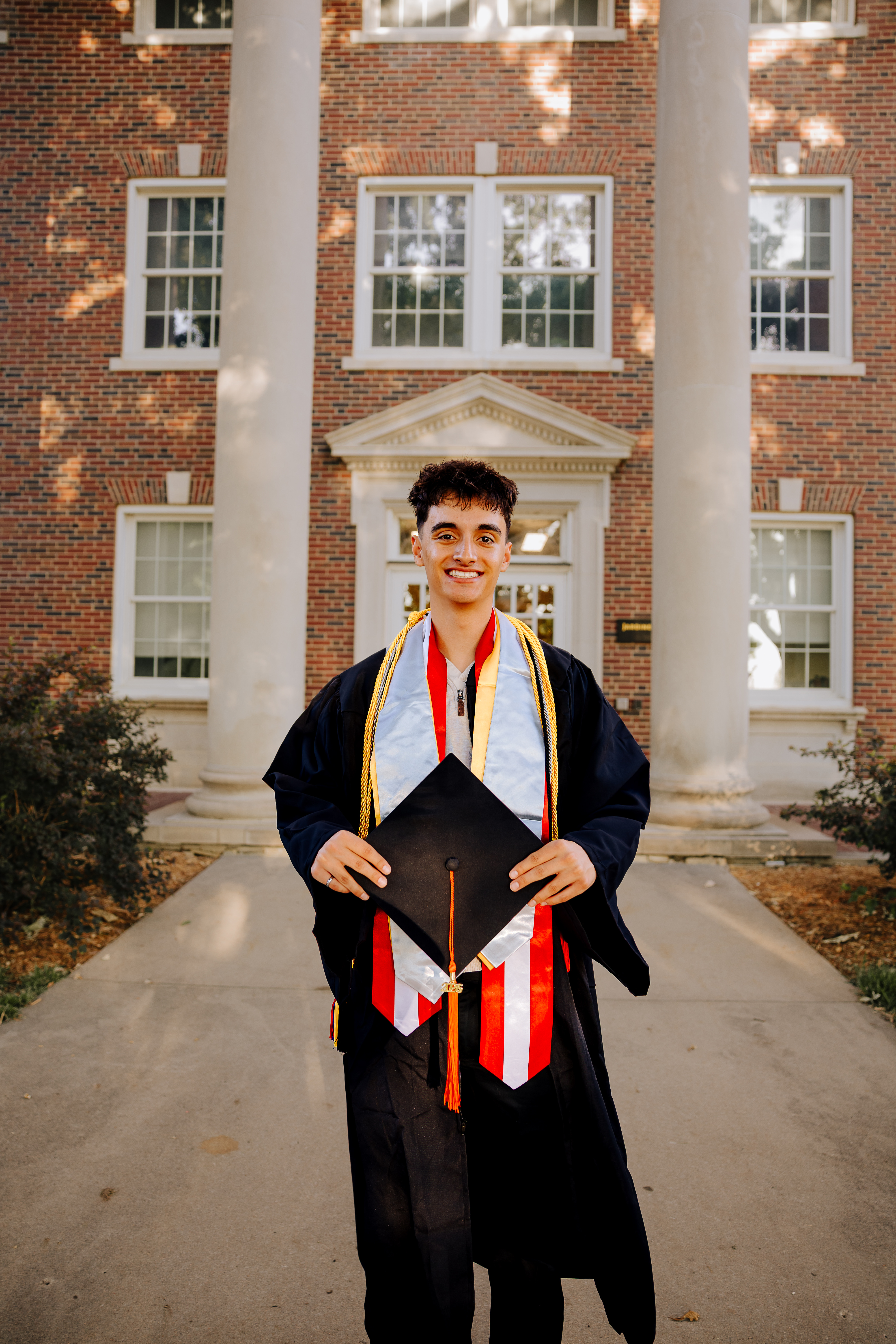 Photo of Michael Boydo standing in front of a Wichita State building wearing their graduation cap and gown, graduation stole, and honors cord.