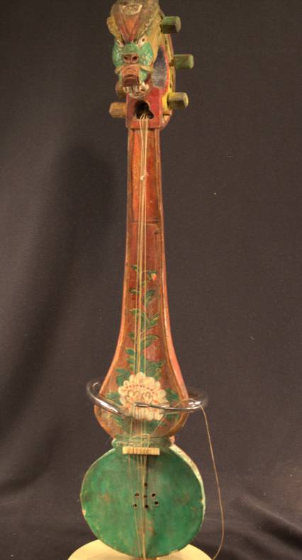 Tungna A string instrument that has a base shaped like an eight. The head of the instrument is carved to look like the head of a dragon. The base is painted green with four gold swastikas. A flower starts at the base of the instrument and makes its way up the neck.