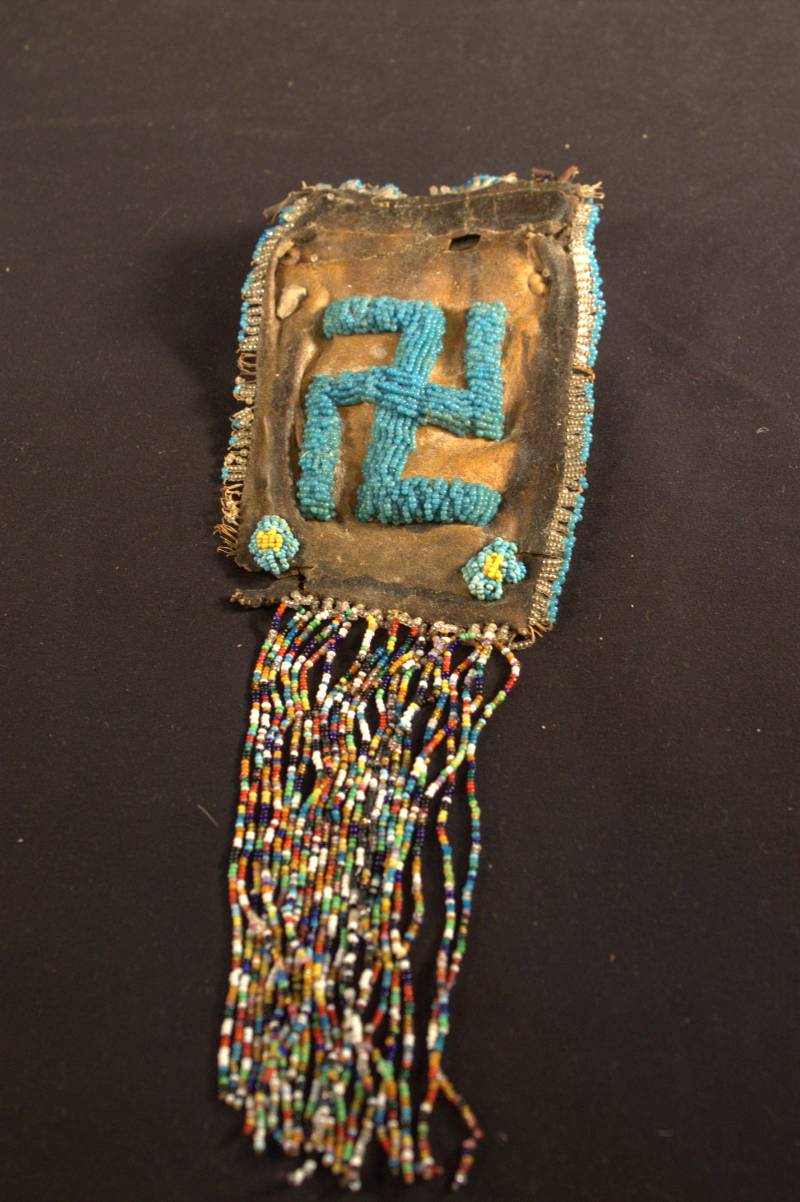 Drawstring Bag A small brown leather drawstring bag that is starting to deteriorate. Multicolored beads hang from the bottom, blue beads create a border around the edge, two flowers made from blue and yellow beads are located in the bottom corners, and in the center is a swastika made of blue beads.