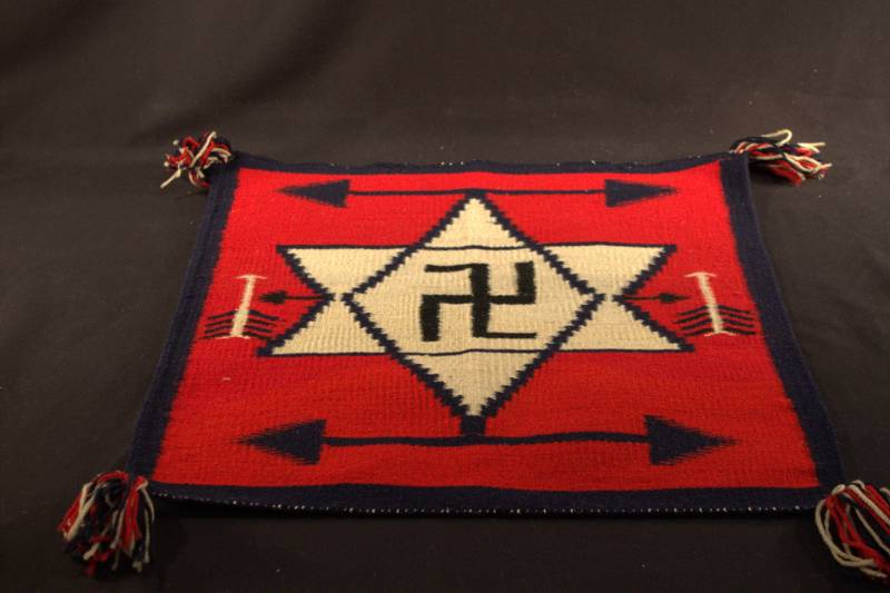 Sampler A square rug that is primarily red with a dark blue border. There are various arrows making a pattern along the edge, and in the center is a white six-point star that has a swastika in the middle.