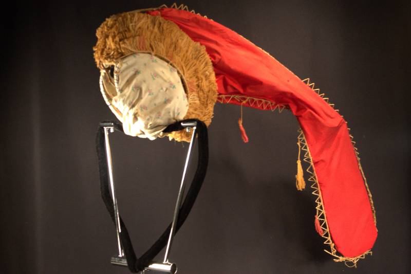 Headdress Headdress is red conical shaped fabric with wooden triangle decoration along perimer. Fabric tassels hanging from conical part headdress part has fibers around outside the fabric covering the hat part is white with pink roses.