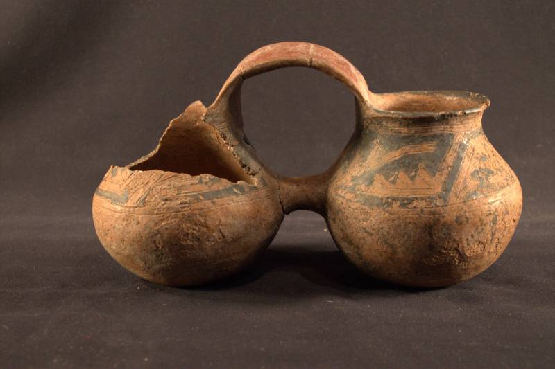Double-Jar Vessel Double-jar vessel is a red/brown color with a black geometric design. Two jars are connected by a handle in the center and the left jar is heavily damaged with the top missing.