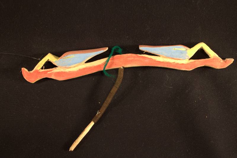 Bird Ornament Long and skinny carved piece of wood with bird-like figures on each side colored red, yellow and blue with a metal spring attached at the bottom.