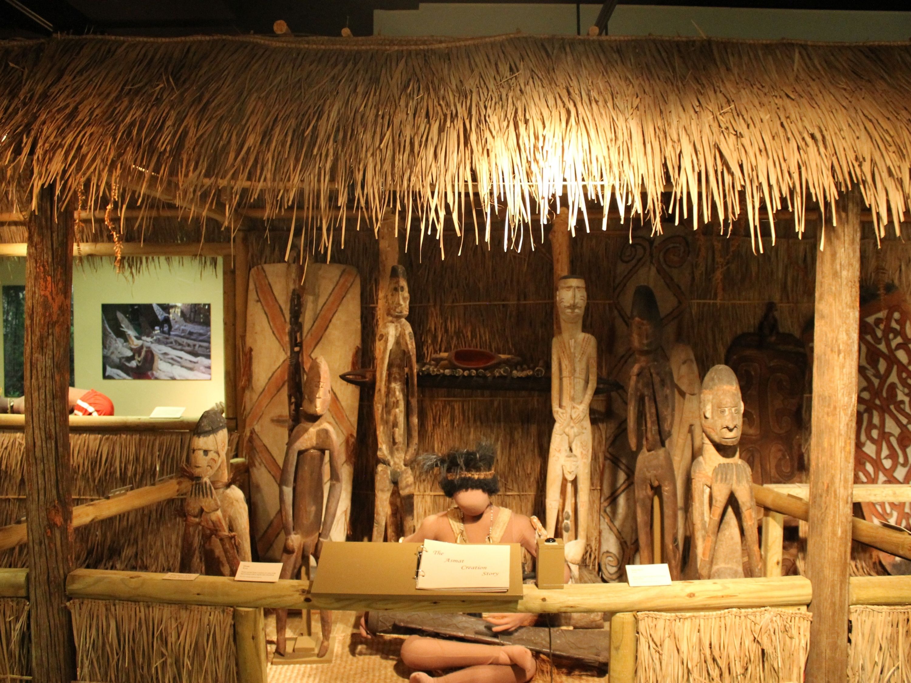 The display depicts a mannequin in an Asmat costume playing a hand drum in a thatched hut. Around him are wooden statues of men. In front is a flipbook about the Asmat creation story.