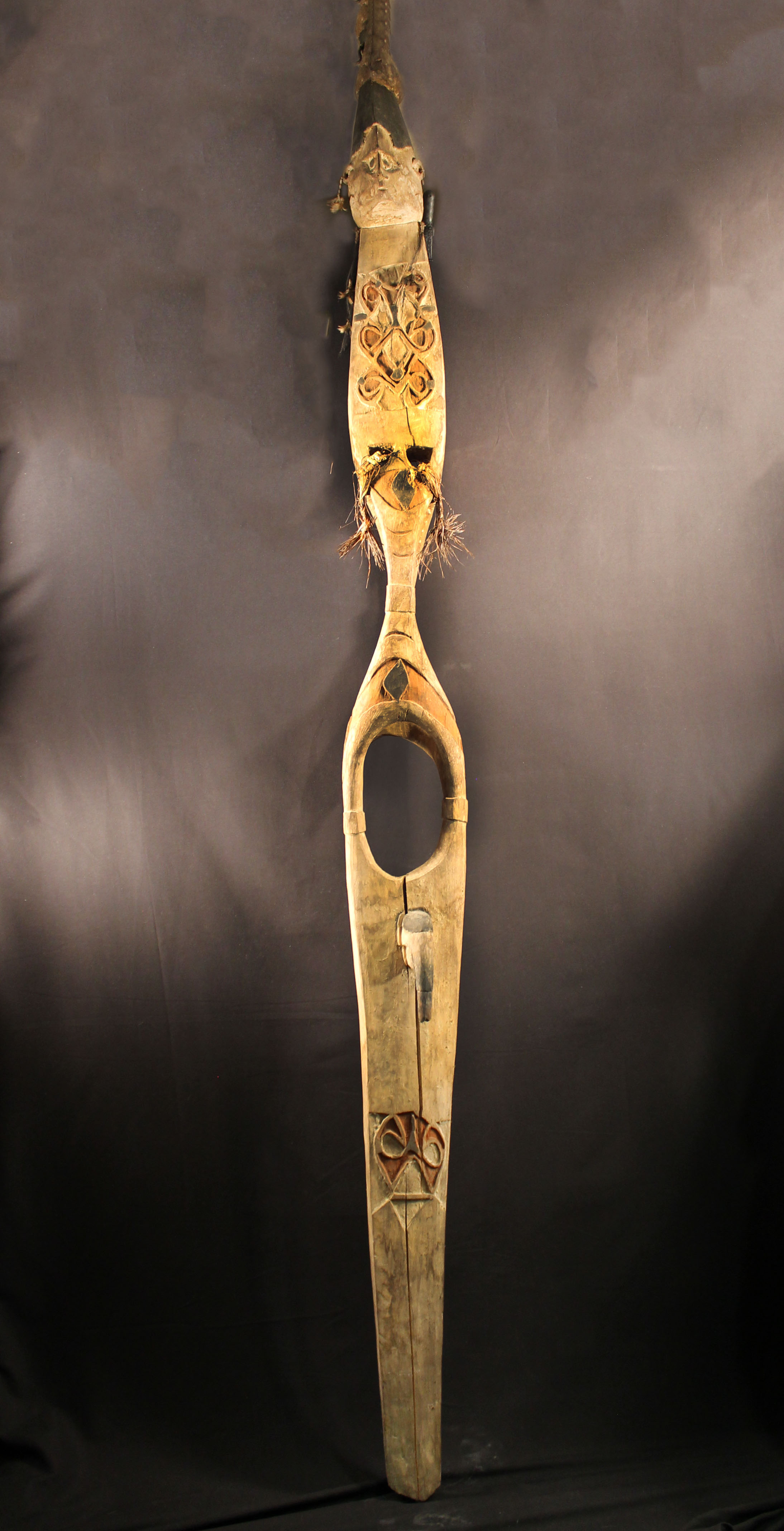 Tall hourglass-shaped wooden carving. The top features an image of a human head with tassels made of leaves, seeds, feathers, and string hanging down. The wood is colored with white, red, and black powders.