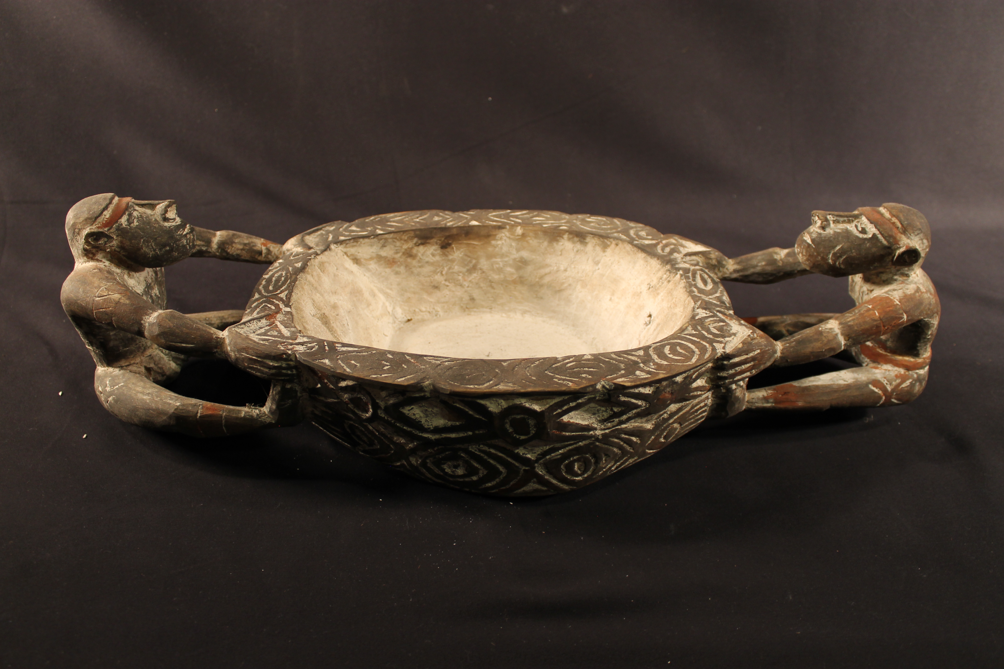 Wooden bowl with white interior. On the sides are male and female figures that act as handles for the bowl. 