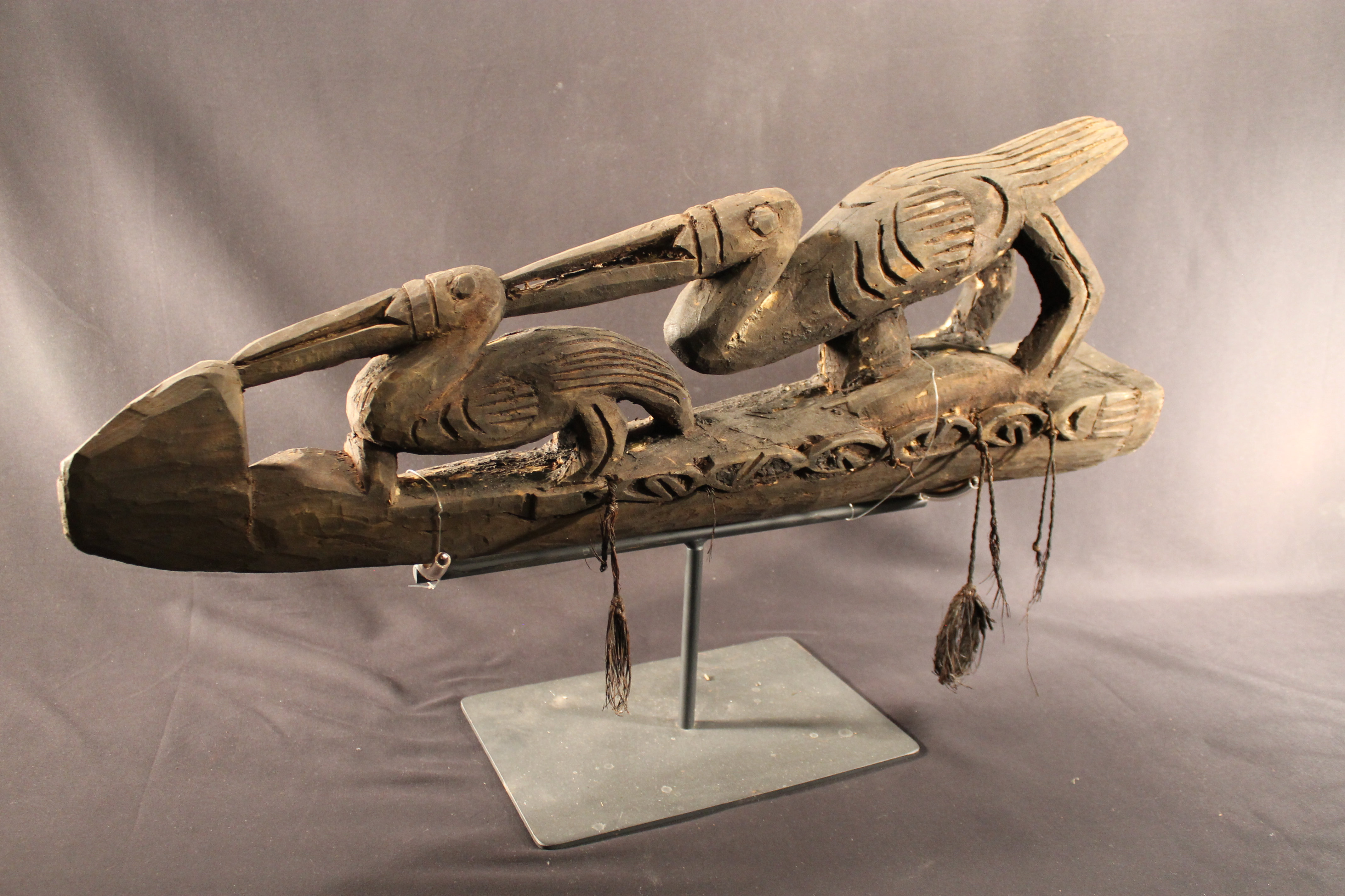 Wood carving depicting two pelicans. Tassels made of plant material connect to the side.