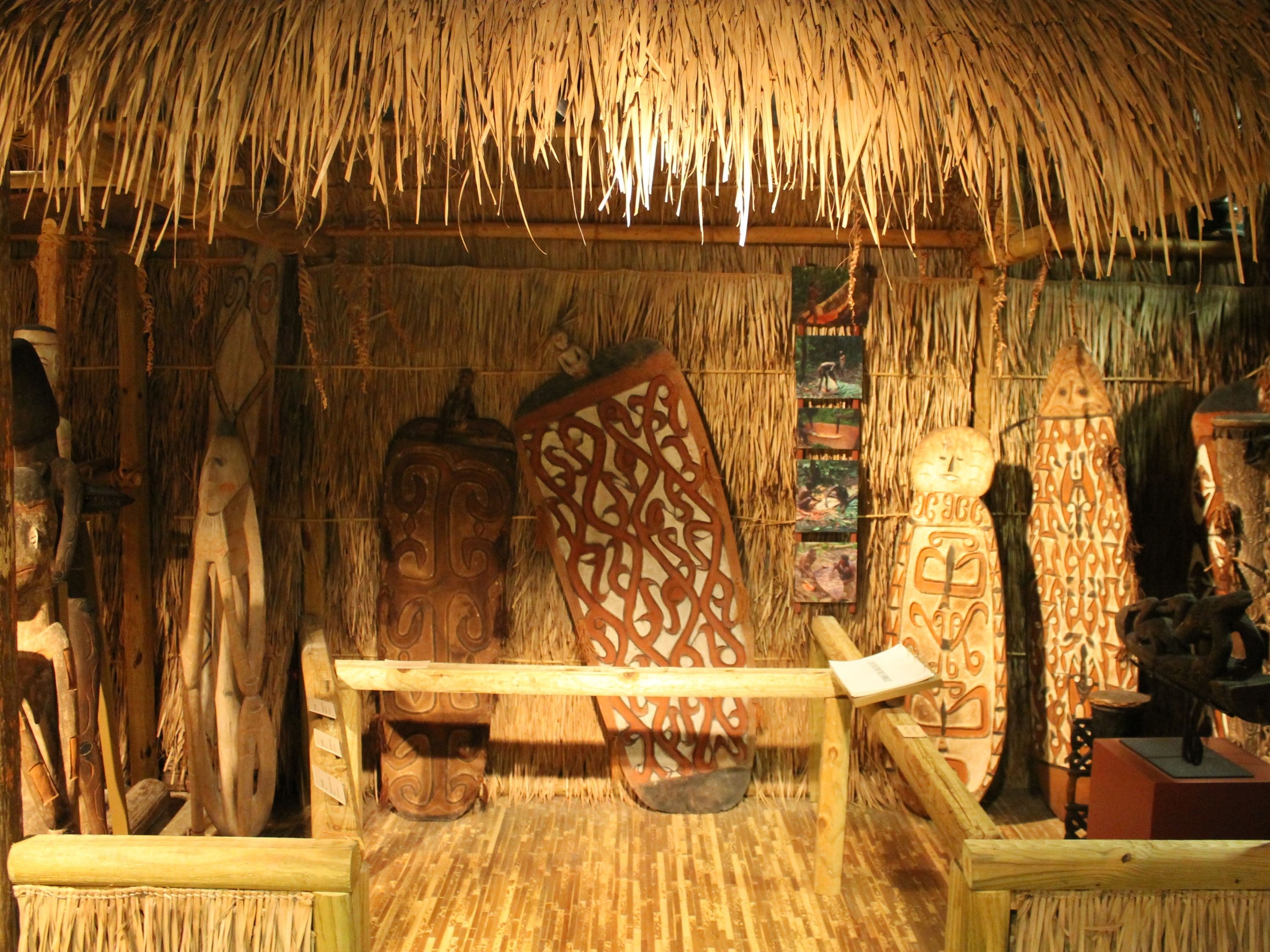 Interior of a hut with shield, drums, and other carvings displayed.  In the front of the exhibit is a flipbook containing information on the display. To the left are five photos that show how these shields are made. The first photo is of a man cutting down the root of a mangrove tree. The second is of two men carving out the removed piece and shaping it with tools. The third is removing the wood’s bark. The fourth and fifth are of the two men drawing on the symbols