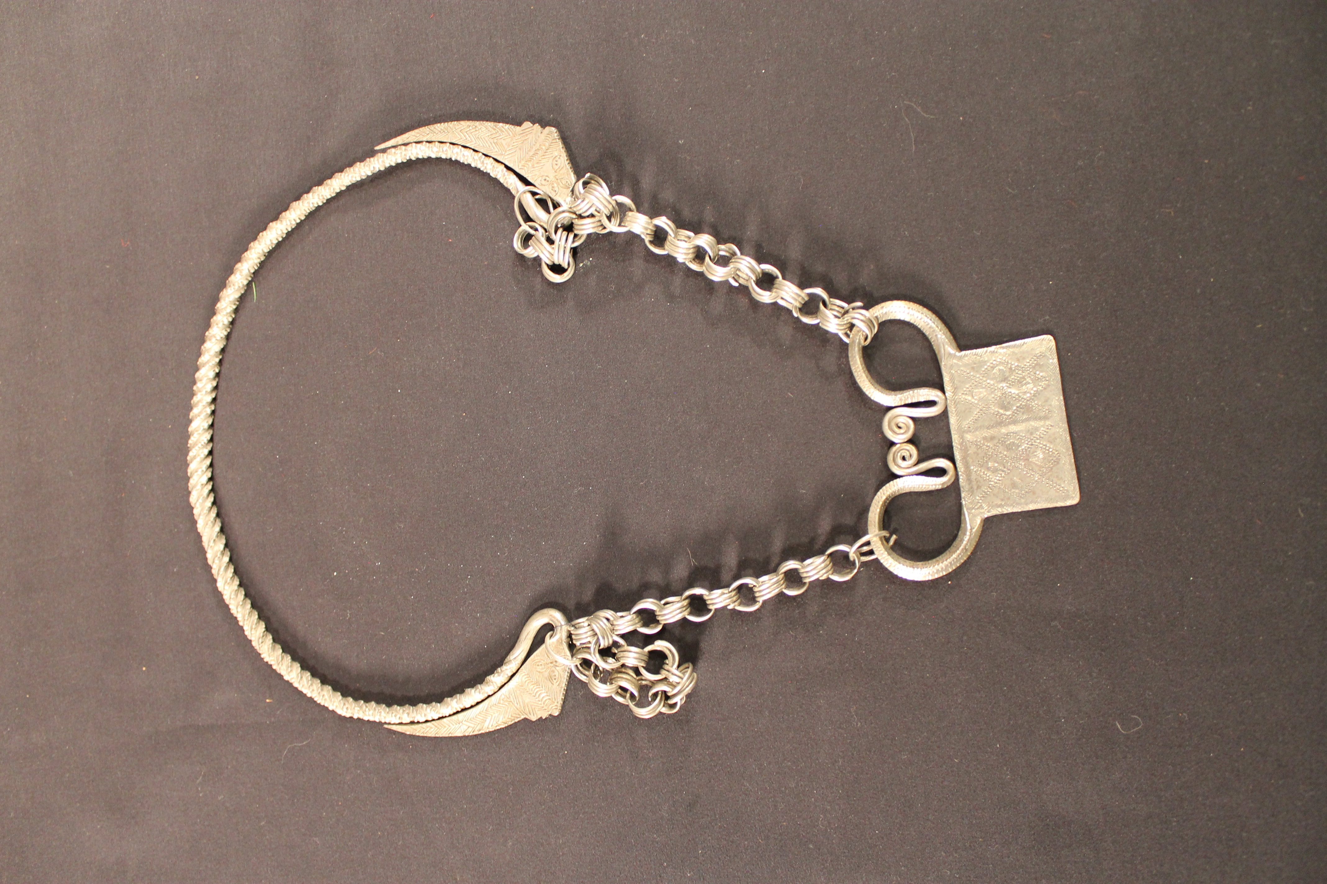 Box-shaped amulet with two large loops connecting it to a linking chain in the middle connecting the amulet's two large loops. Around the back of the necklace is a rope of silver metal. 