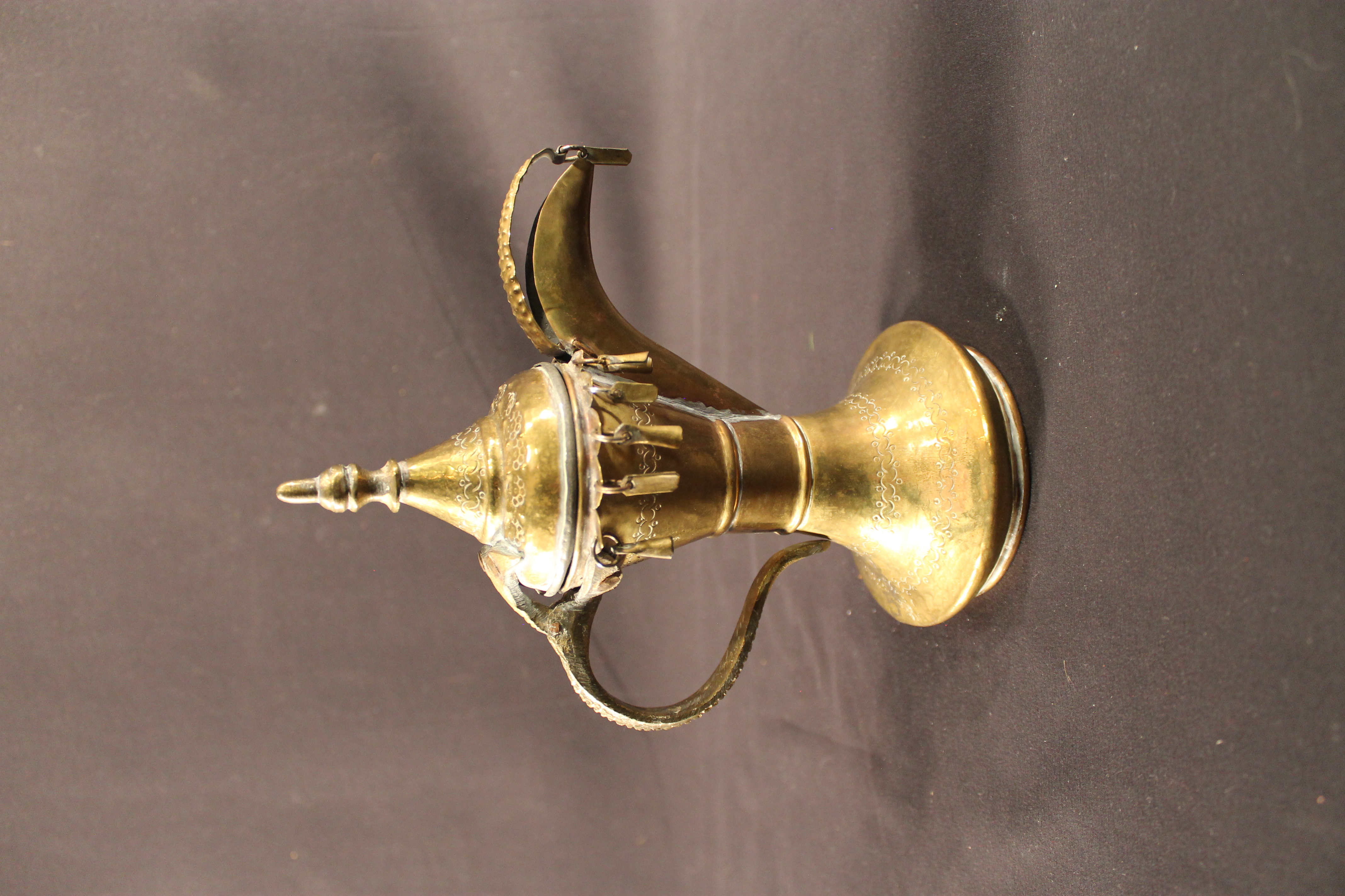 Brass coffee pot featuring an ornately designed hammer into the metal. Various shaped charms hang off the pots on the bottom of the lid and the end of the pot’s spout. 
