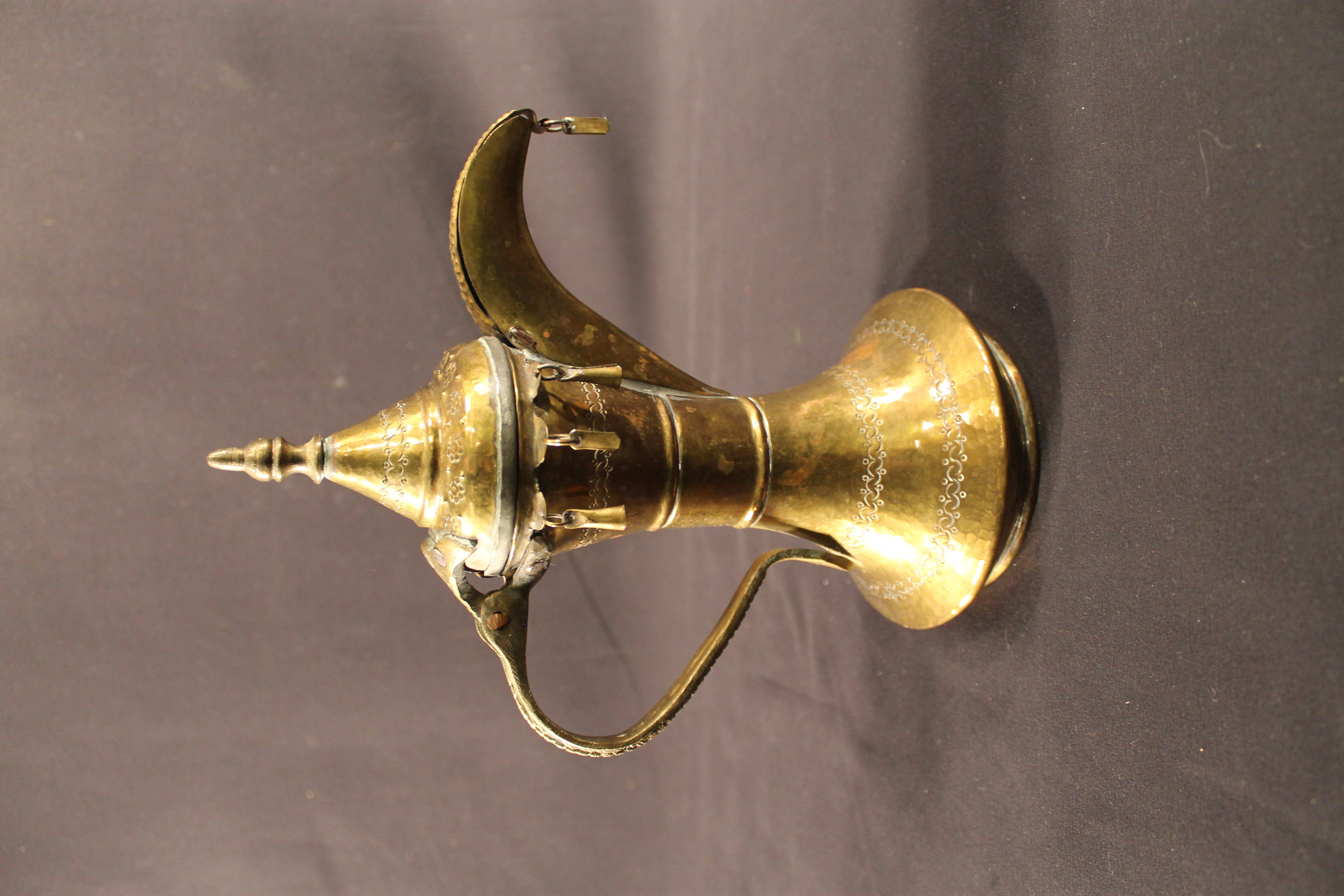 Brass coffee pot featuring an ornately designed hammer into the metal. Various shaped charms hang off the pots on the bottom of the lid and the end of the pot’s spout. 