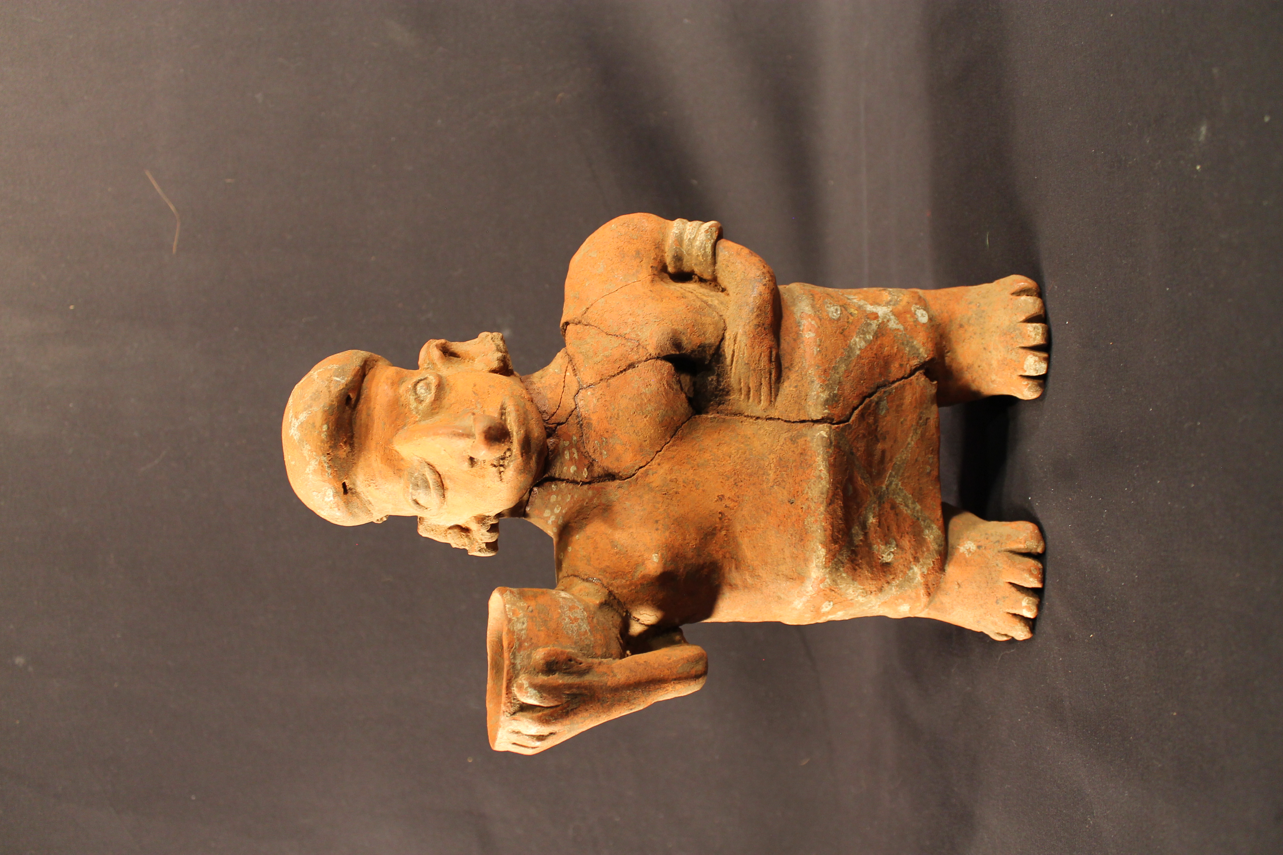 Orange male figure that has a large chest and lower body. Additional designs that have been painted white have been faded. They are wearing a garment that is decorated with a crossing pattern. They also wear jewelry pieces on their left forearm and a nose ring. In their right hand is a decorated pot. The statue is in poor condition, with a large portion on the right-hand side being broken and repaired. 