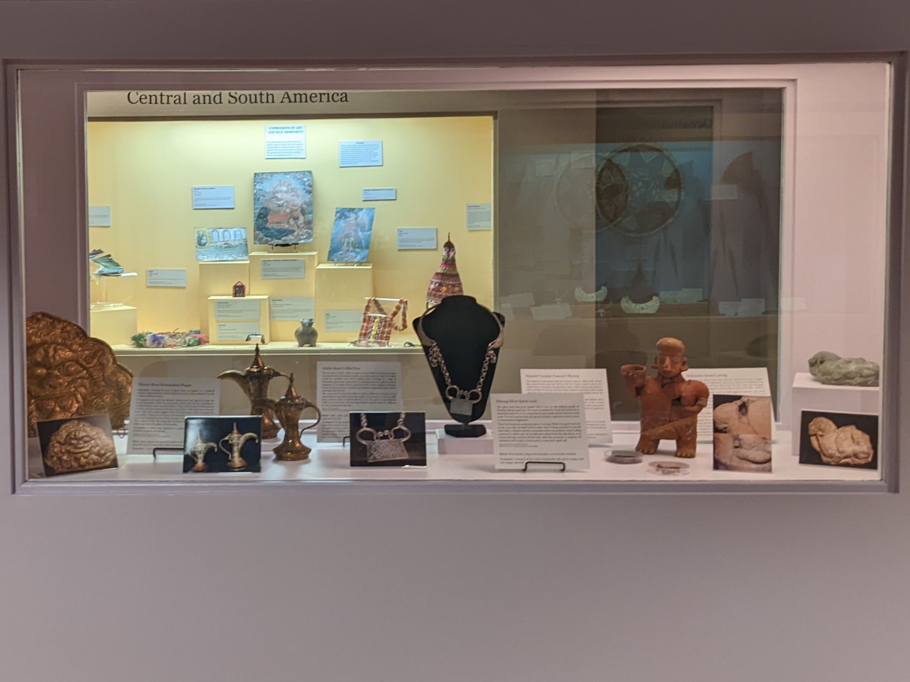 The center cases hold five restored items, including a brass plaque, brass coffee pots, silver necklace, terracotta color ceramic figure, and a stone carving with a green tint. Besides the objects are label information on the object and before treatment photographs.