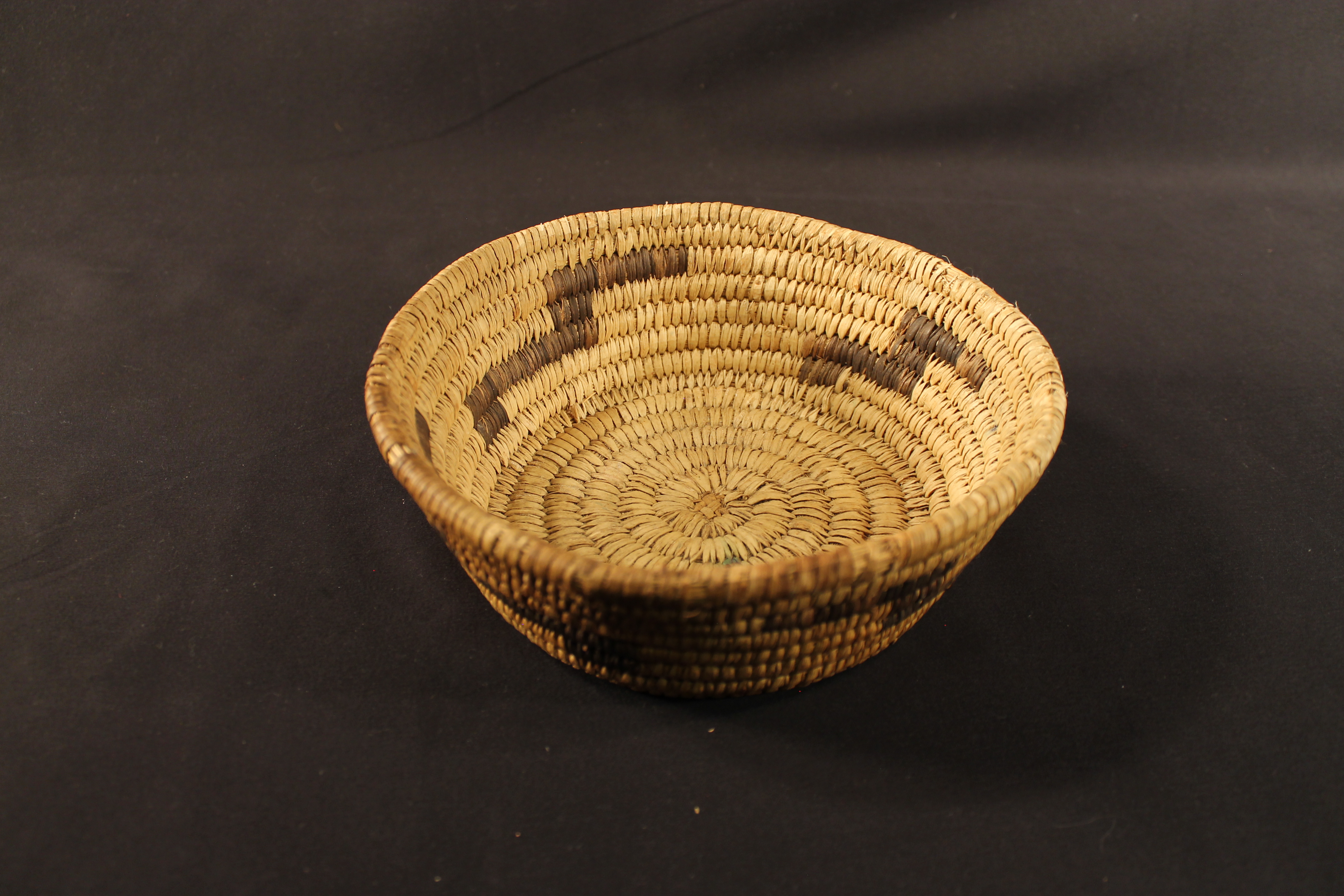 Brown woven coiled basket made up of several rows, and depicts an “s” like pattern made of black woven reed. 