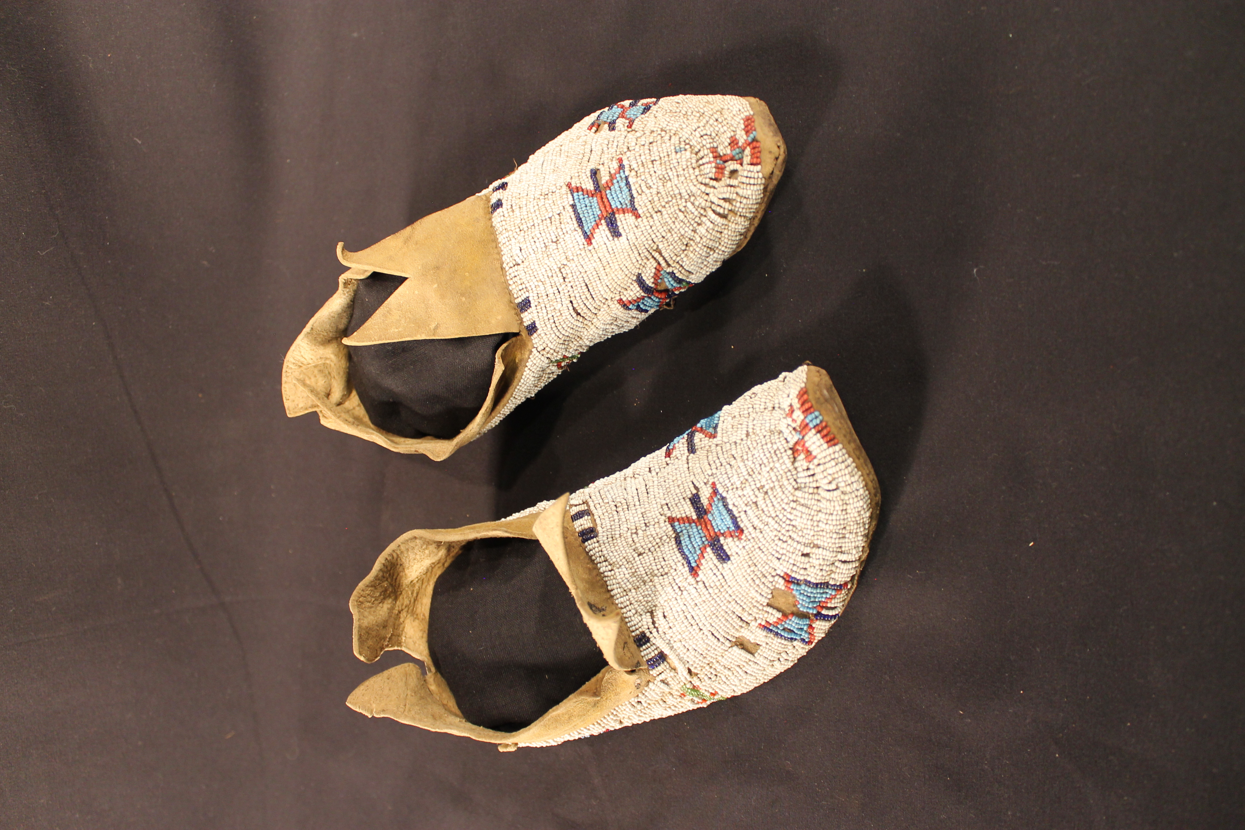 Beaded shoes with a pattern featuring two blue triangles outlined with red beads connected together with a black beaded bar. The remainder of the design is filled in with white beads. The upper portion of the shoe is exposed tan hide. 