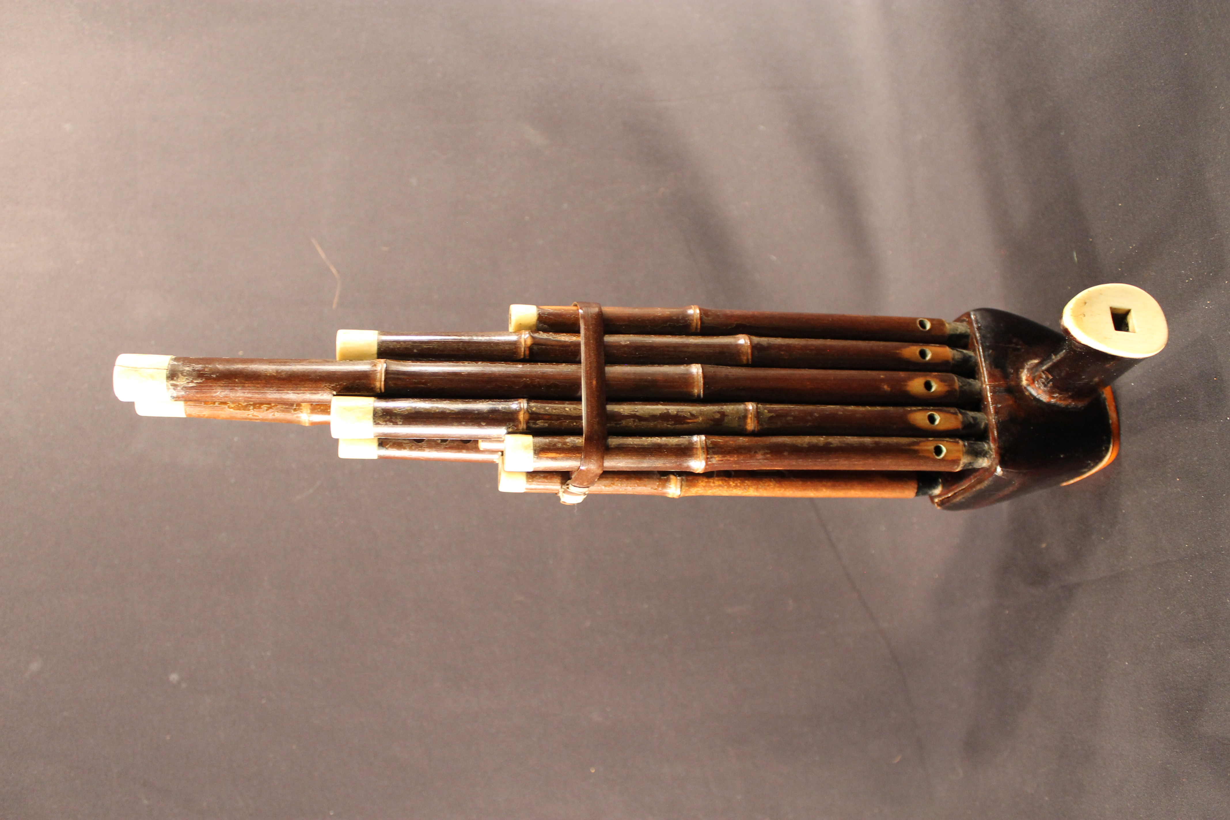  An instrument made up of ten dark wooden pipes and a mouthpiece. The pipes are held together with a wooden band, then they connect to the mouthpiece that extends off of the side. Ivory pieces surround the top of the pipes and on the mouthpiece. 