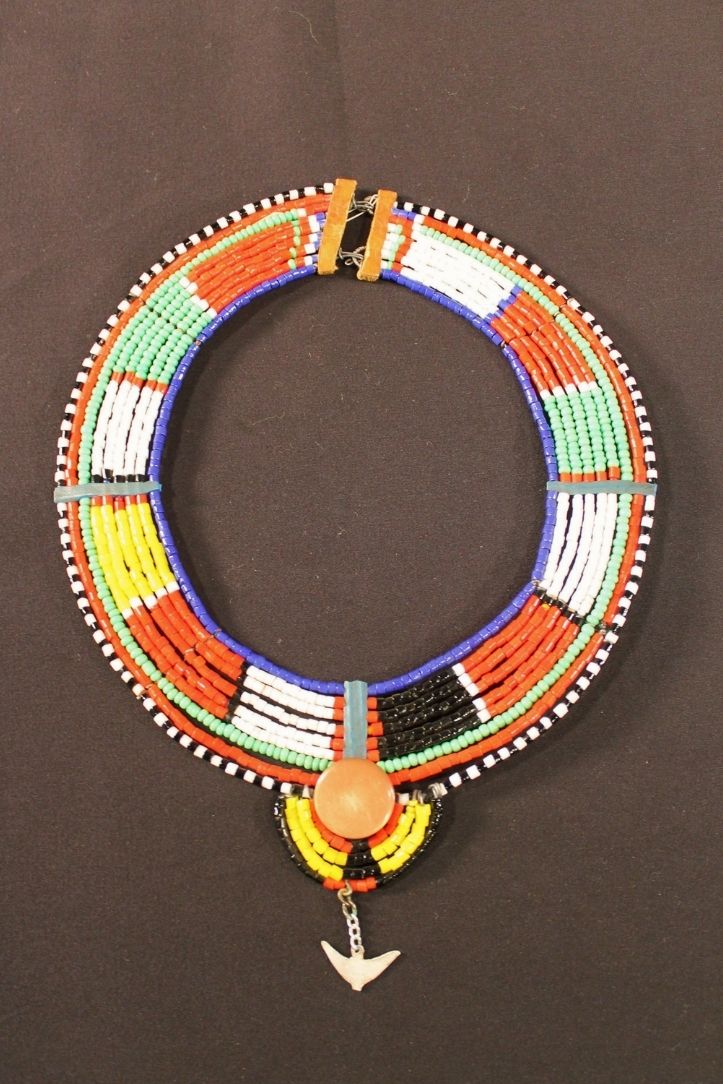 Circular beaded necklace with blue, white, green, yellow, red, and black beads but has a plastic button and a metal bird dangling from the bottom of the necklace.