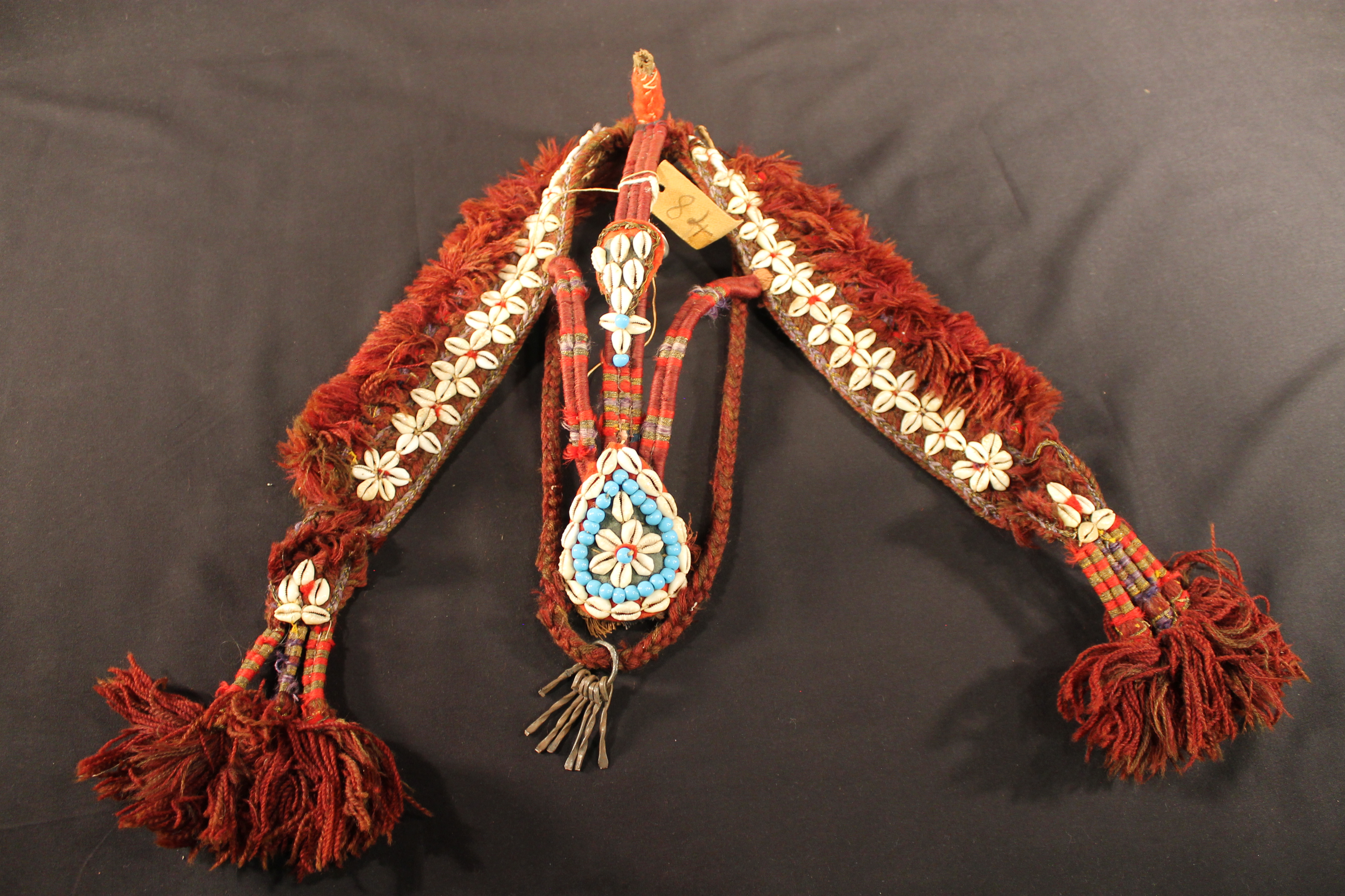 Braided yarm horse headgear has woven in shells and beads to form a star-shaped design on the side. In the center is a shell and bead pendant in the shape of a teardrop. 