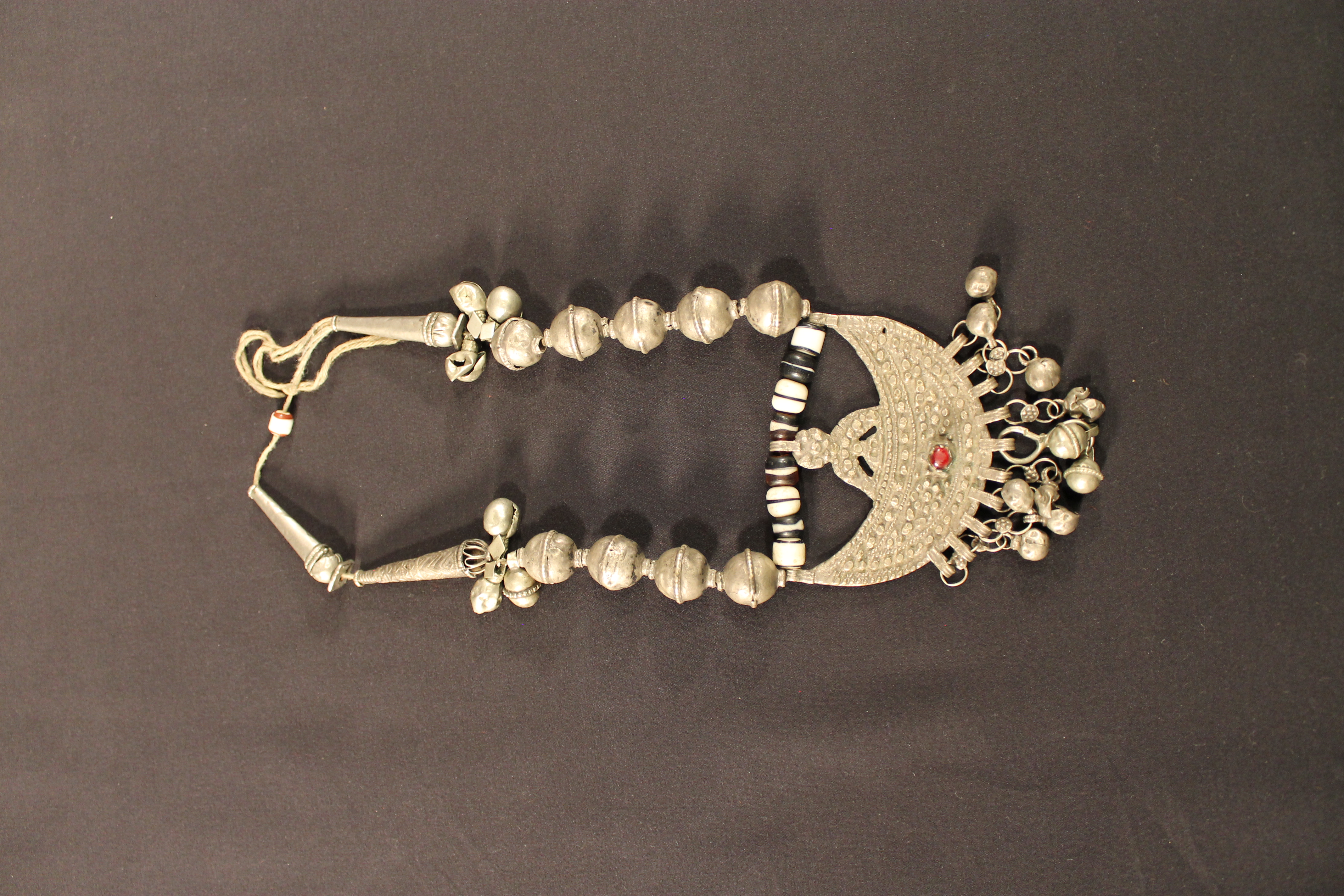 A silver multi-shaped necklace has a crescent pendant with a red gem embedded in the center with bells on either side.