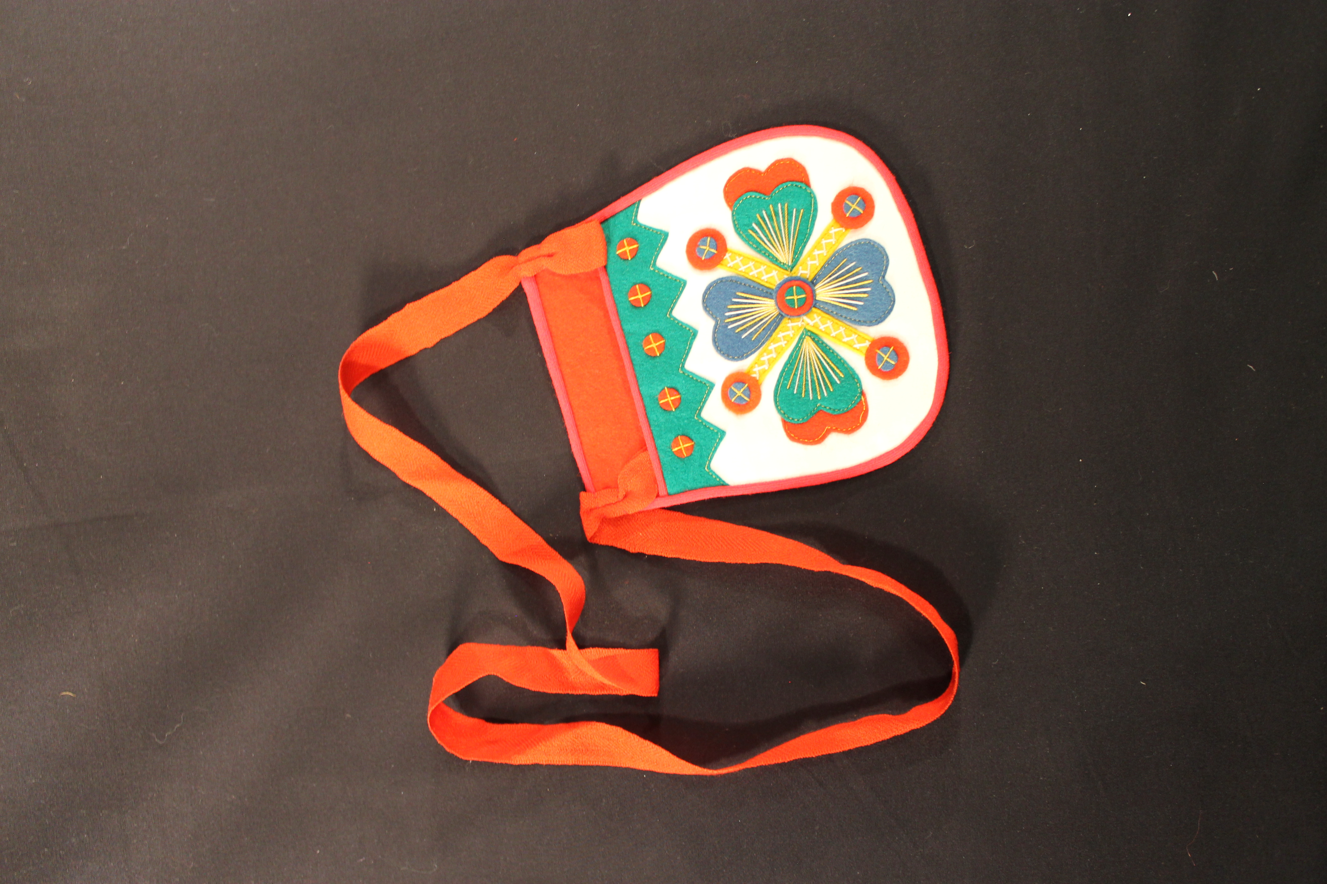 Pocket-shaped felt purse with a red back and ribbon strap. On the front is an applique of floral and heart-shaped designs.