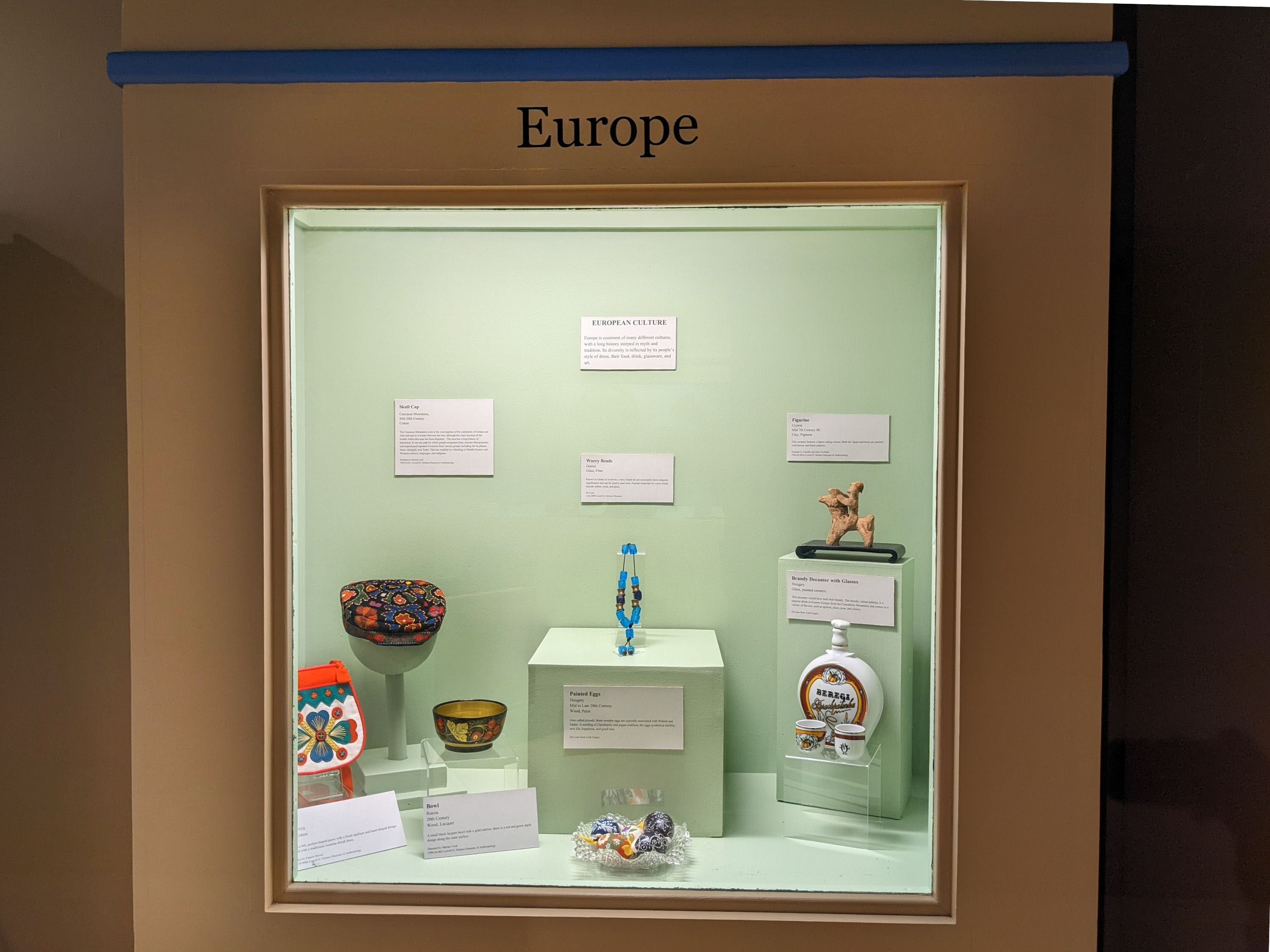 Case with green background. Objects in the case are a bag, hat, bowl, beads, painted eggs, a horse figurine, and a decanter with glasses. Label attached to back wall and bottom of the case. 