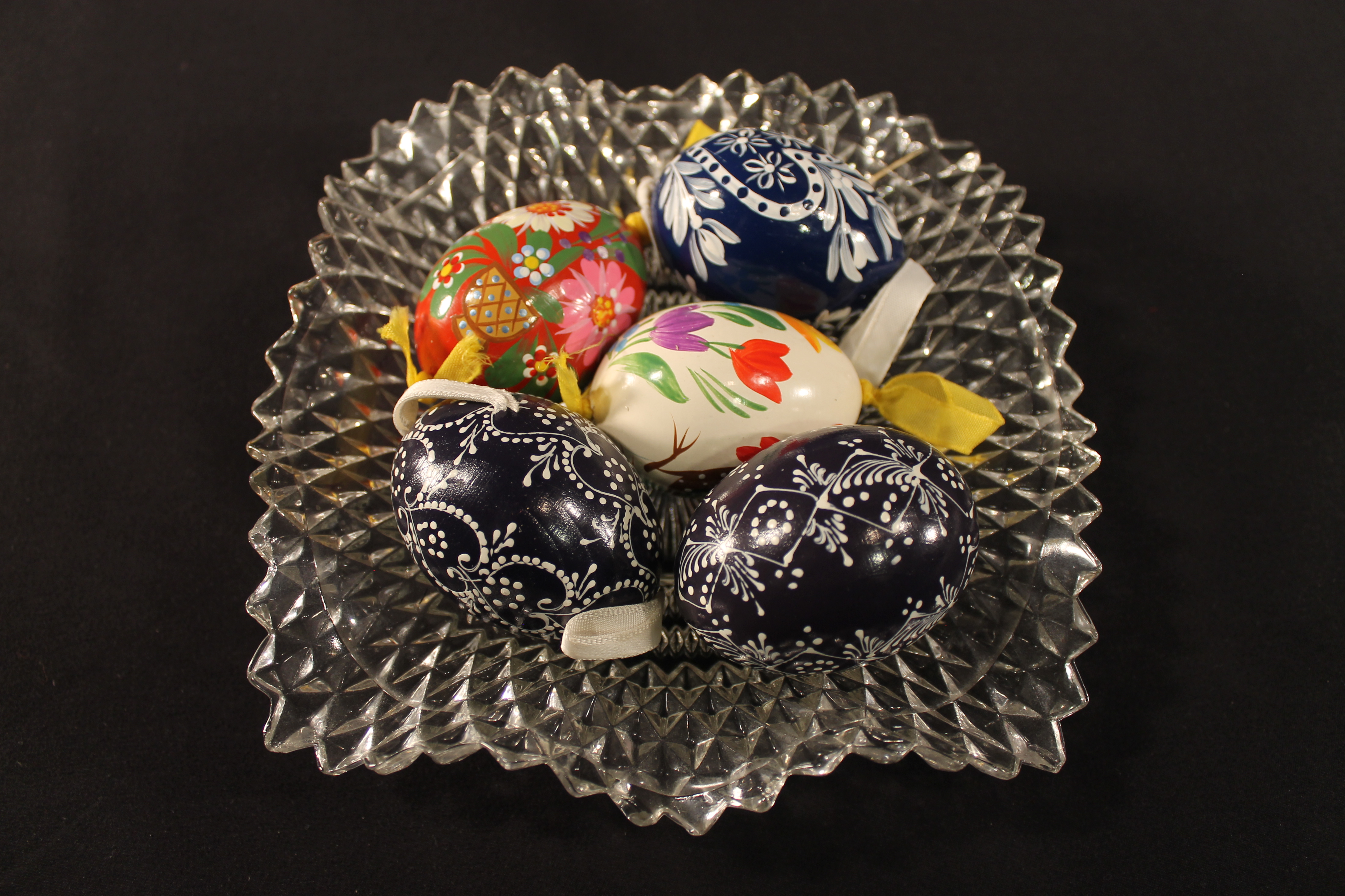 Five painted eggs. Three of them are blue and white with ornate designs with a white attached ribbon. One has multi-colored tulips and a yellow ribbon. The last one is red with various colors painted on it. 