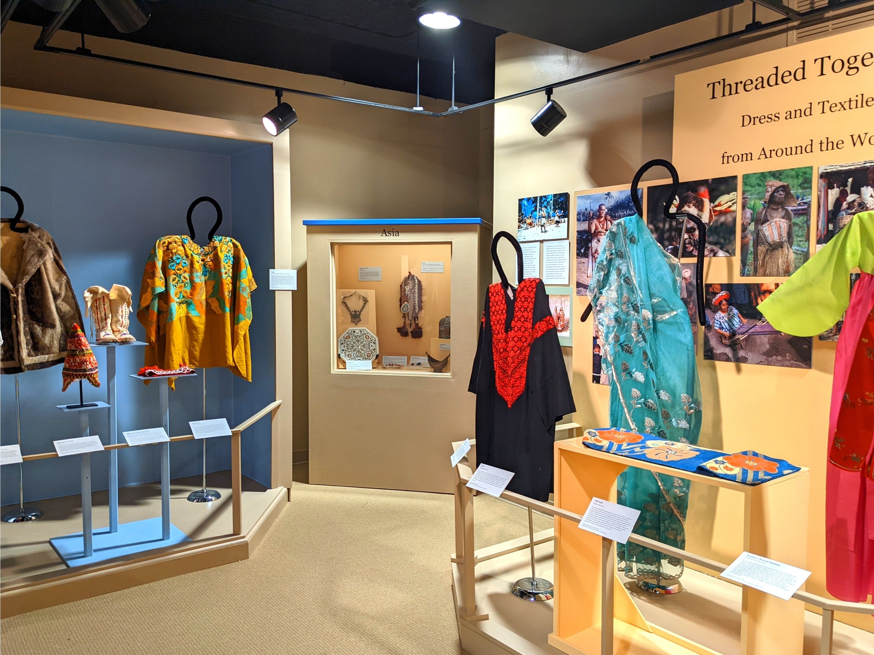 Three displays in a museum gallery. Two of them feature clothing from around the world on a mannequin, while the other is a case with objects in it