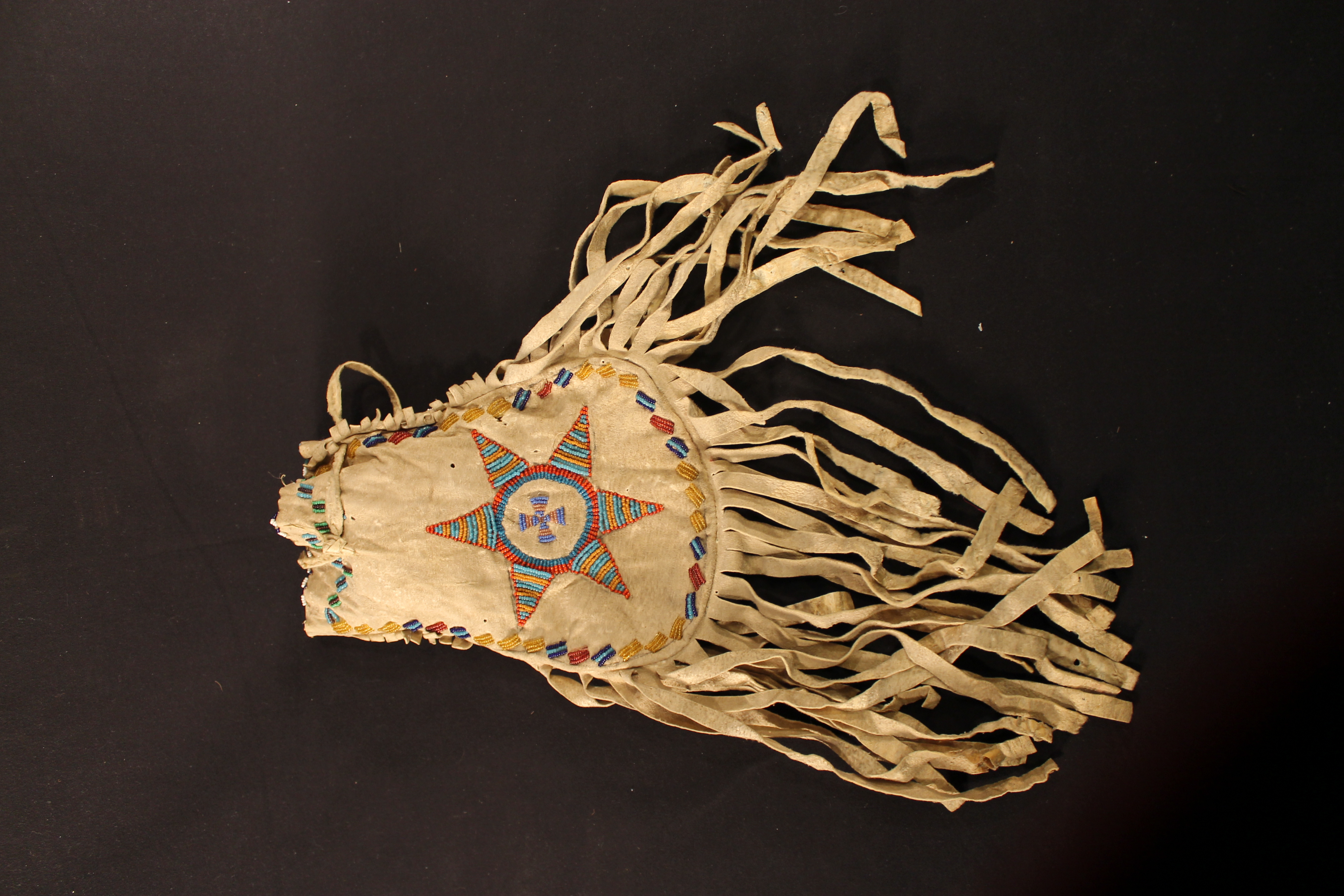  The deerskin bag is decorated with a large six-pointed star of blue, gold, and orange beads, with a cross of blue and pink beads in the center. The bottom half is lined with fringe.