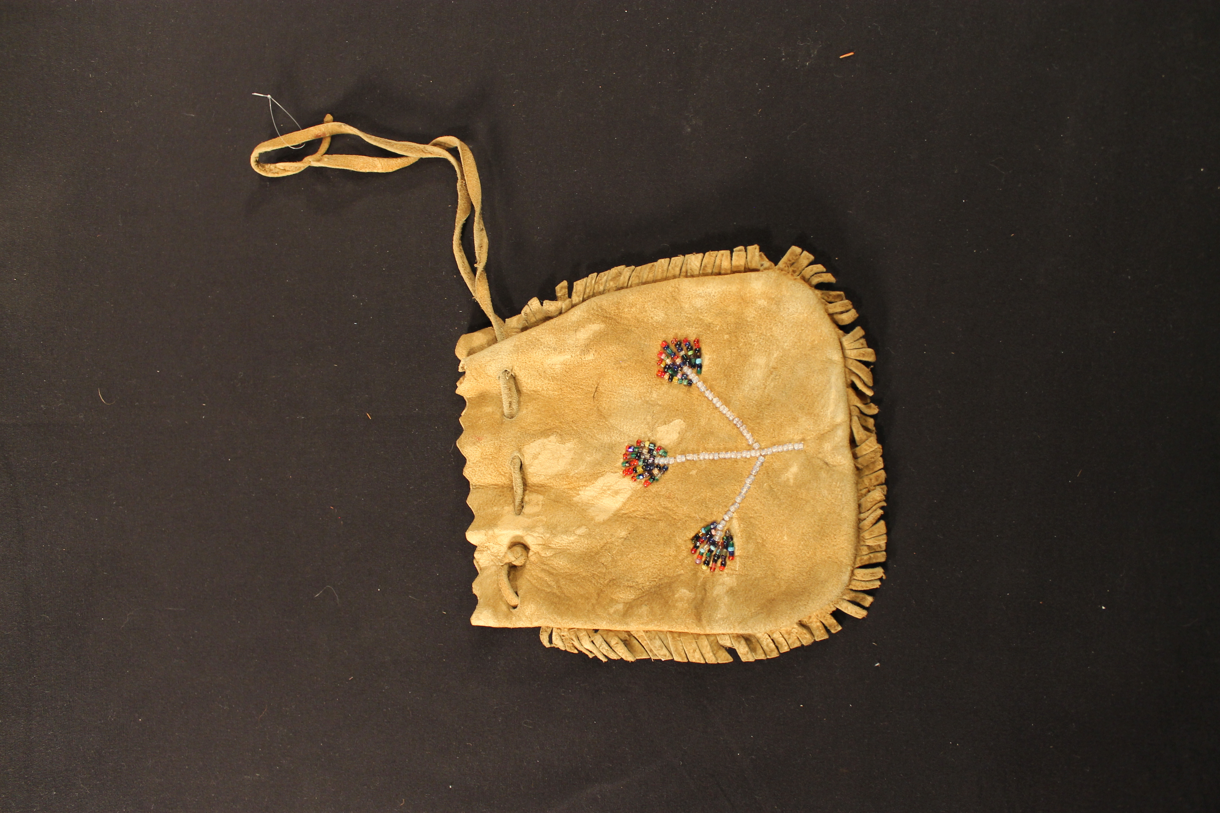 Bag made out of tan leather with multi-flower seed beads on each side. The bag has a drawstring and there is fringe that goes around the bag.