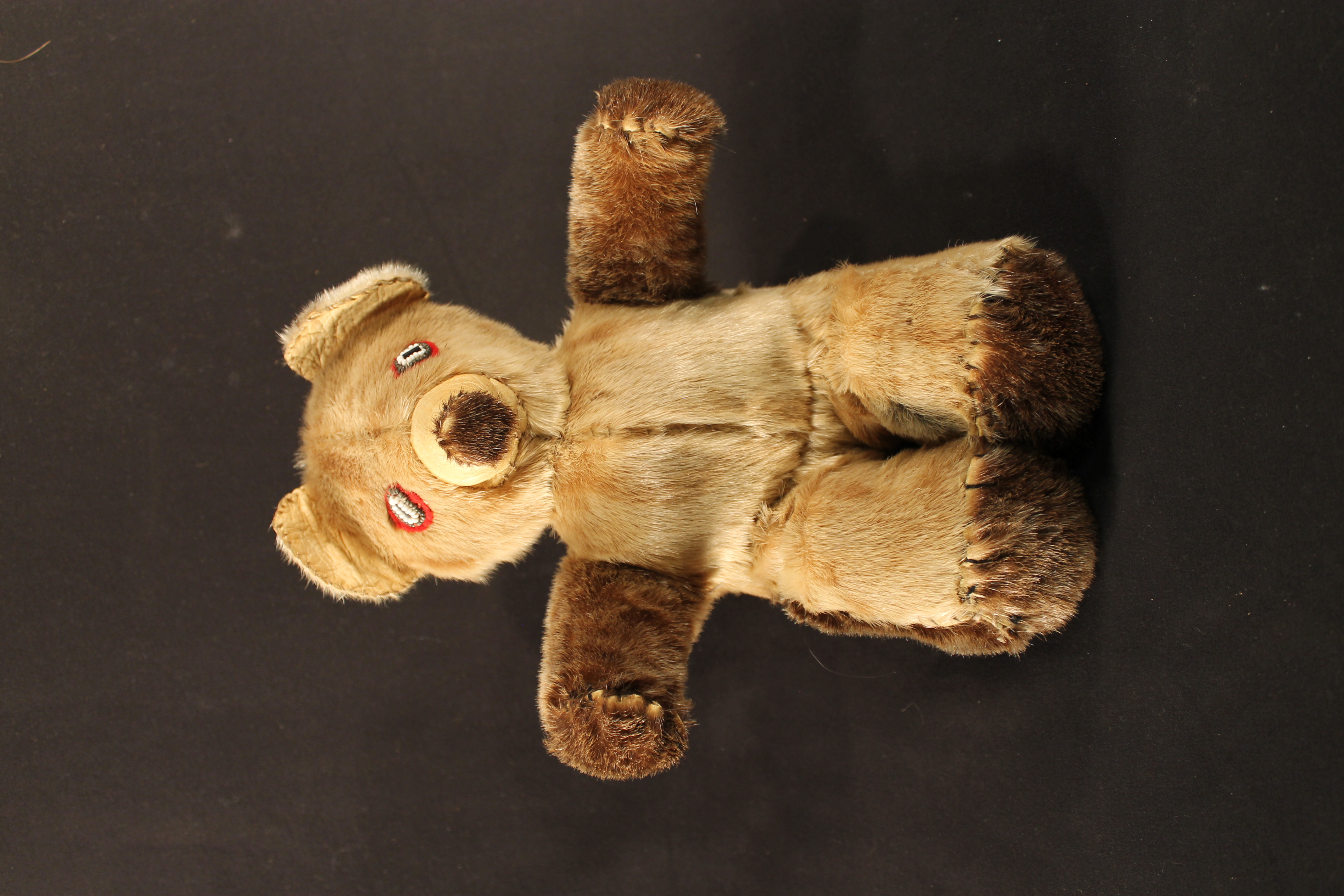 Teddy bear made of fur that is both sandy and chestnut brown. It has a fur body with eyes made of red felt with black, white, and gray beading.
