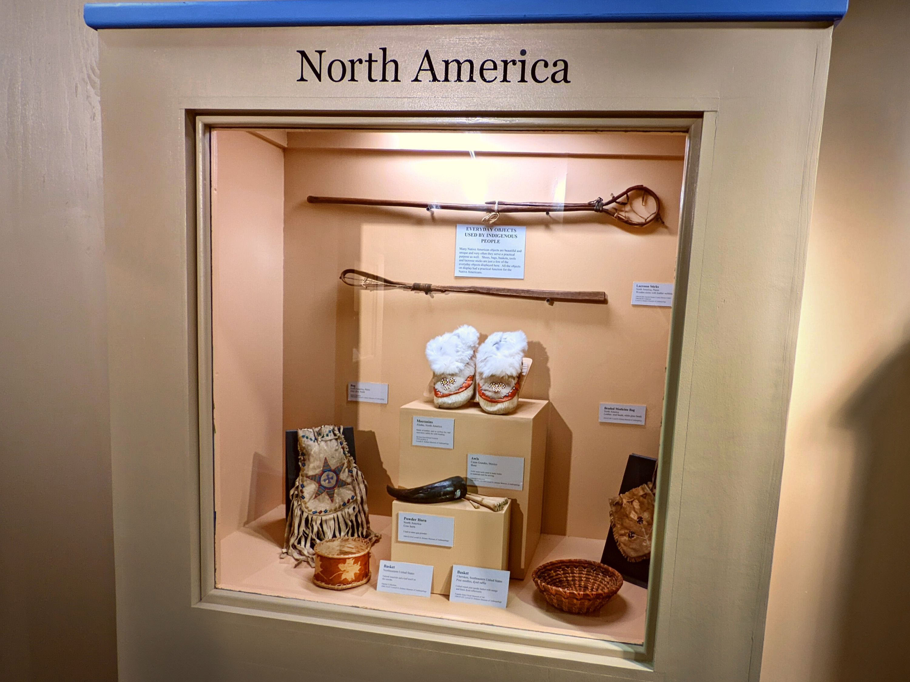 Case with an orange background with objects mounted on walls and on display on the surface of the case. Objects inside include two lacrosse sticks, two beaded pouches, two baskets, moccasins, awls, and a powder horn. Label information for objects on both the back wall and on the bottom surface. 