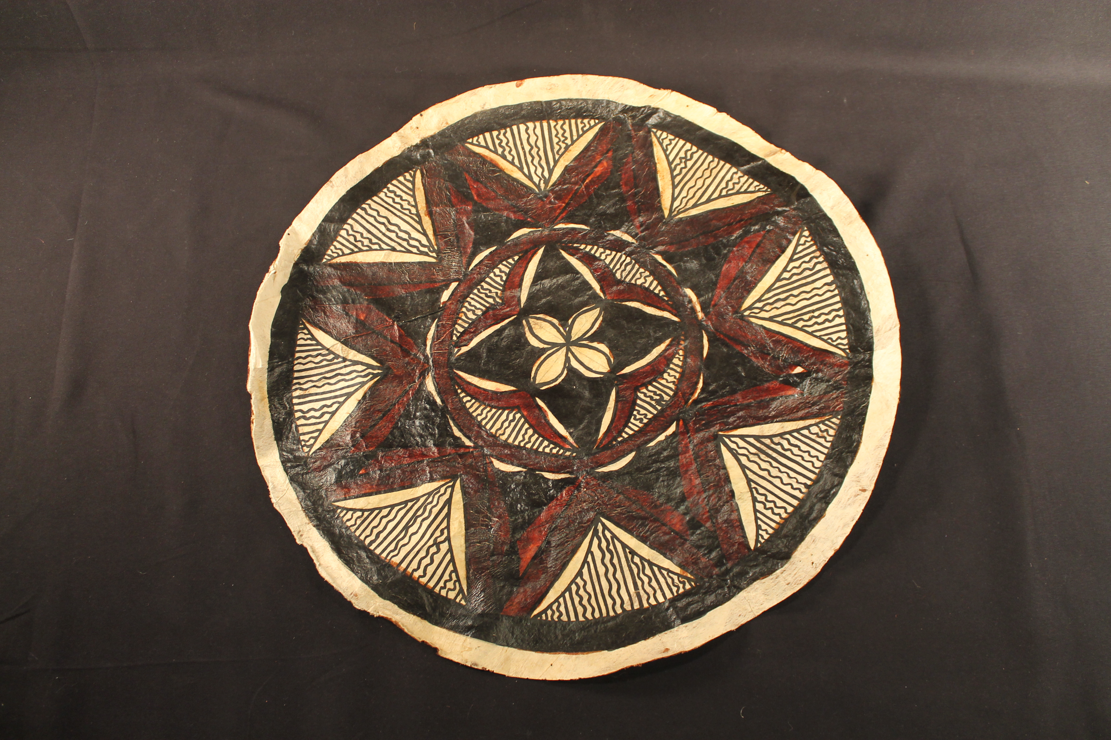 Circular bark cloth made of flattened wood. The bark has a black, red, and maroon-colored design on a tan background. Black straight and squiggly lines radiate out from designs. 