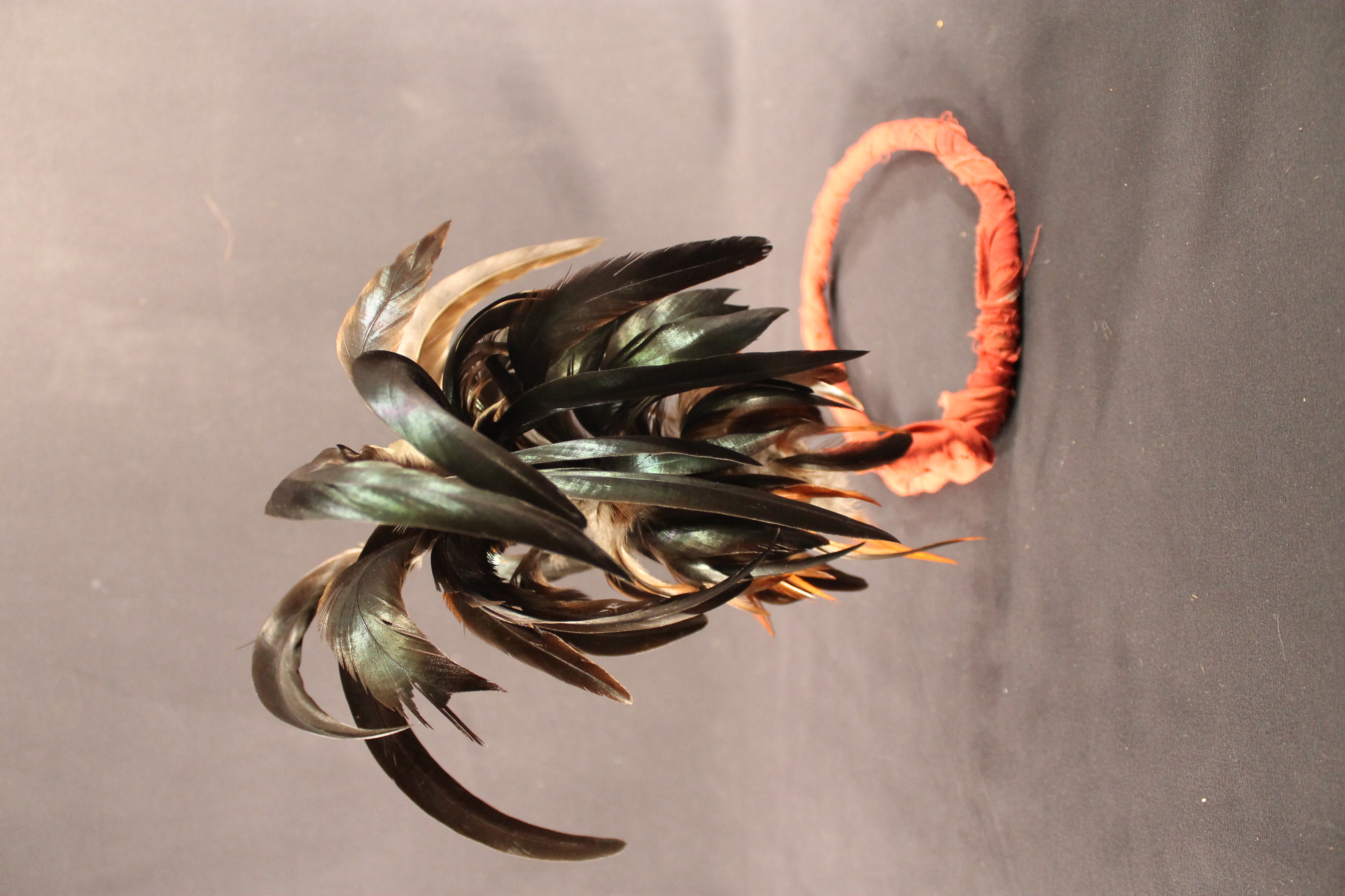 Headdress with metal base wrapped in a red cloth decorated with iridescent, green, and brown feathers on top.