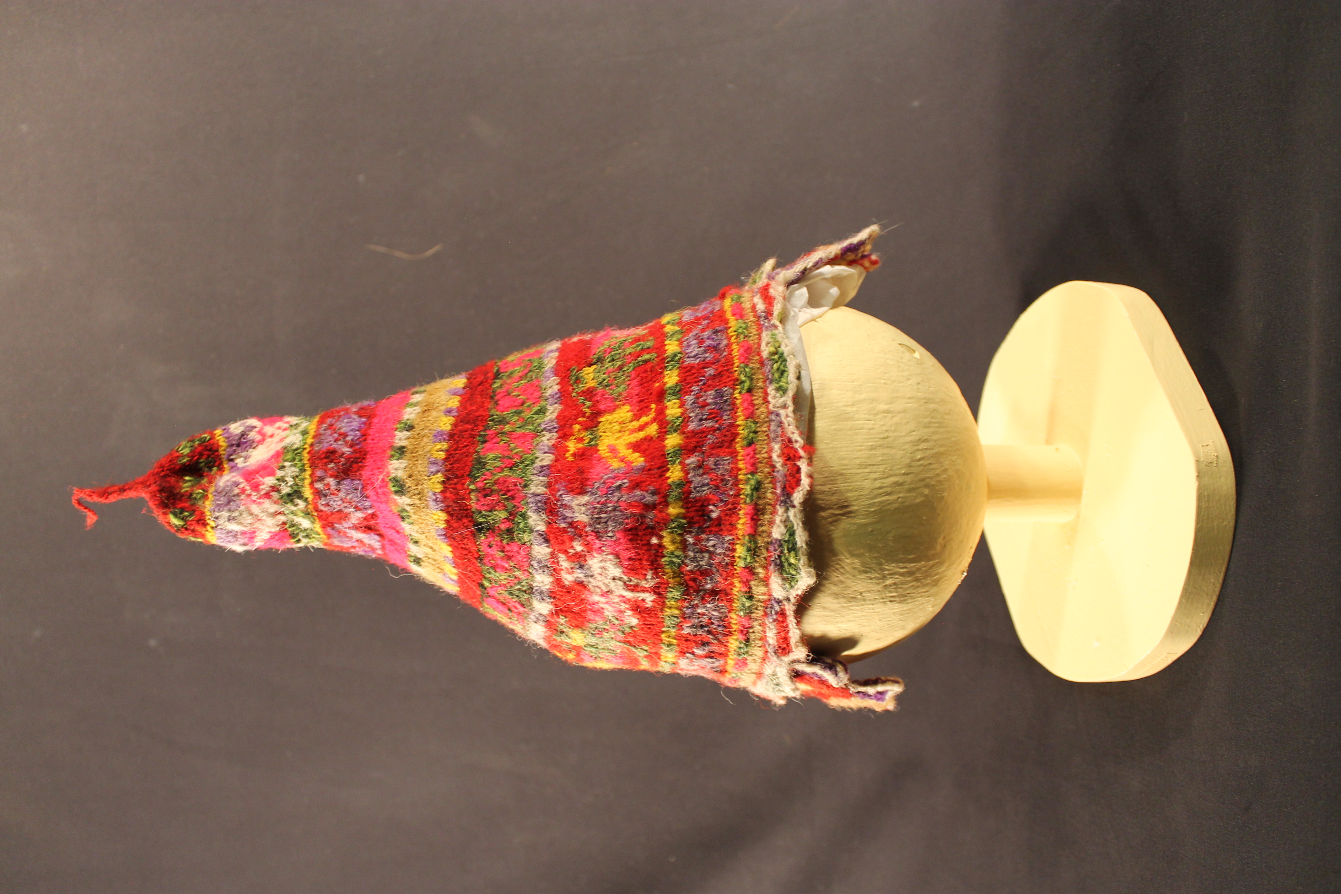 The cap is made of red, green, yellow, purple, white, pink, and tan wool. The cap has two flaps on either side and there is a string that stands straight up on the top.