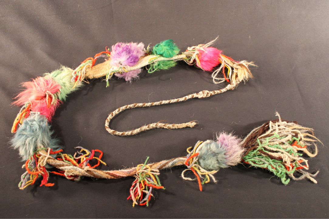 The sash has many colored puff balls of purple, green, pink, and blue. The ropes of the sash are made up of cream, gray, brown, and tan woven fibers. 