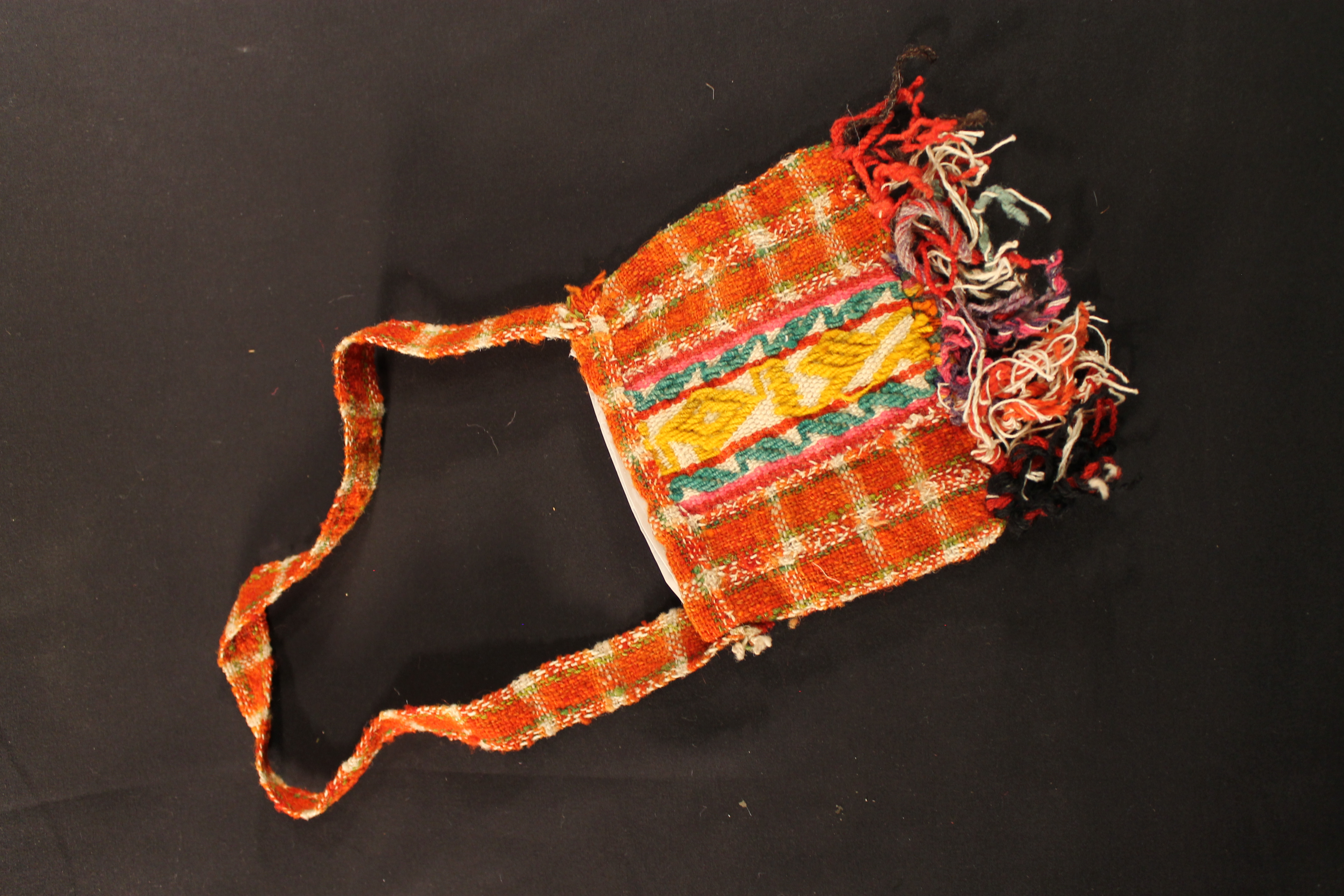 Small, woven wool coca bag. The bag is mostly orange-red with aqua, yellow and pink accents on the central design. Multicolor fringes at the bottom.