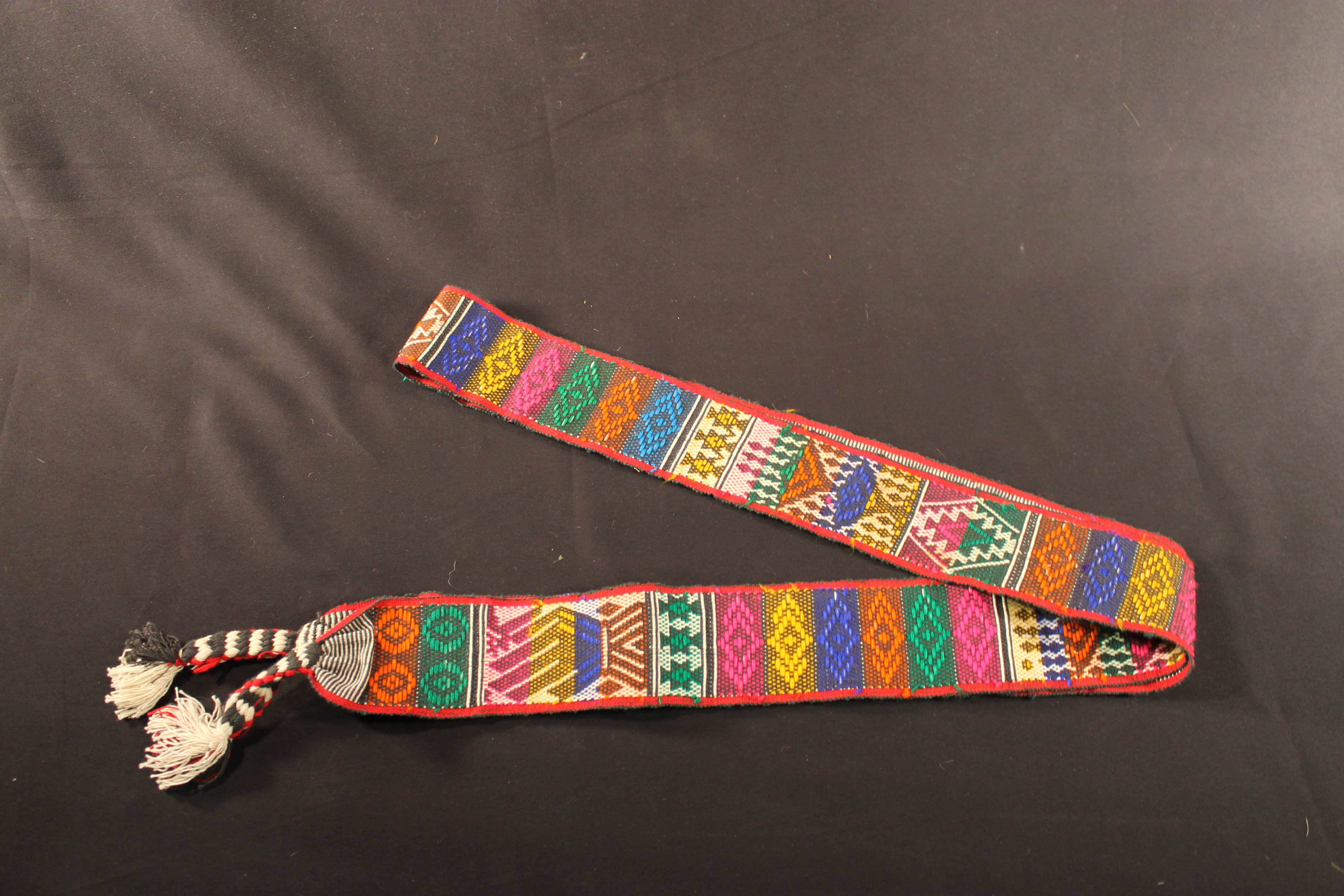 Woven sash that shows designs consisting of geometric shapes with depictions of animal and human figures. Red, pink, yellow, green, orange, blue, aqua threads are used throughout designs. On either side of the sash are braided tassels