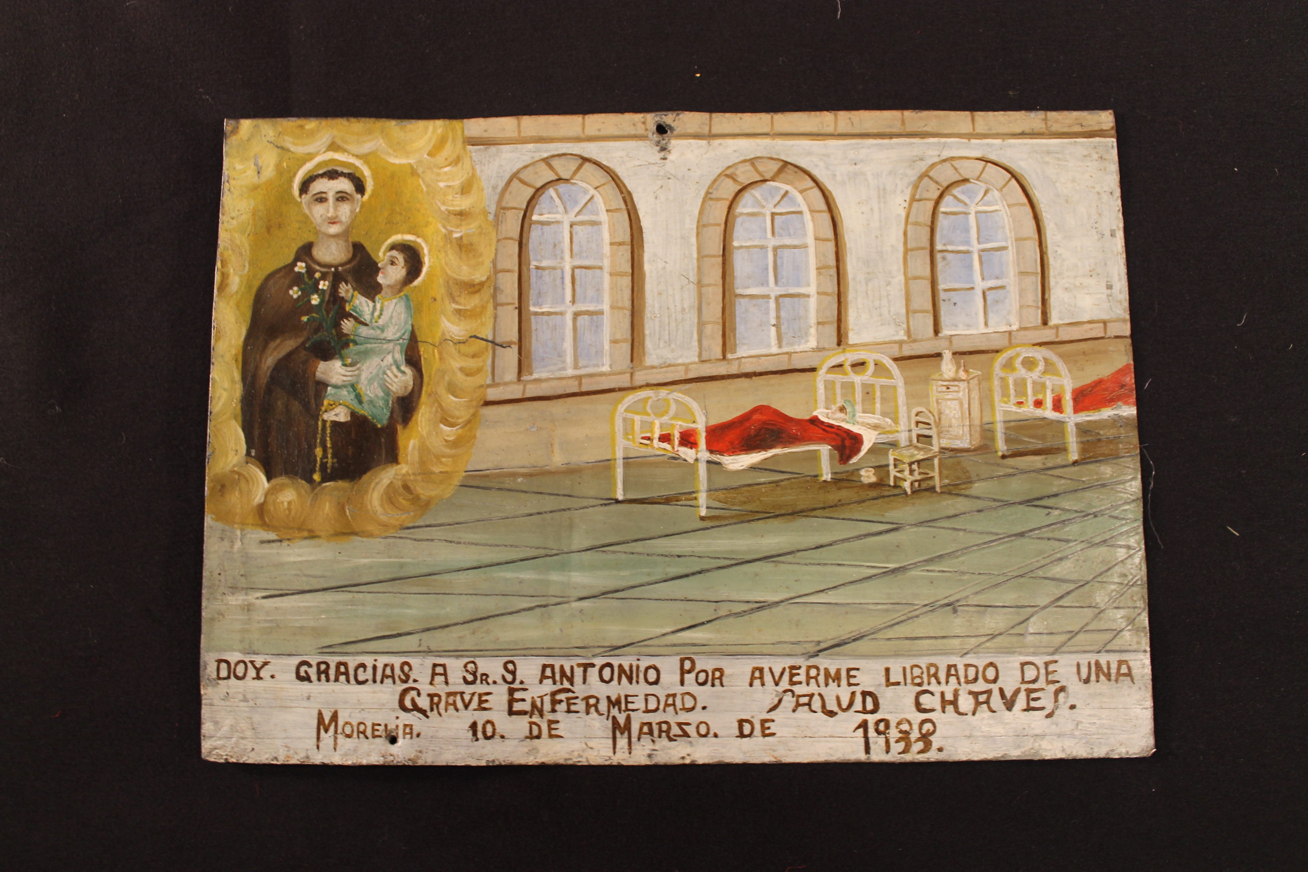 Retablo painting on tin of a patient in a hospital. Saint and baby on the left top corner. Text on objects reads  "Doy Gracias a Sr S. Antonio por averme librado de una grave enfermedad. Salud Chaves. Morelia 10 de Marzo de 1933” English translation: I thank Saint Antonio for having freed me from a serious illness. Cheers Chavez. Morelia March 10, 1988”
