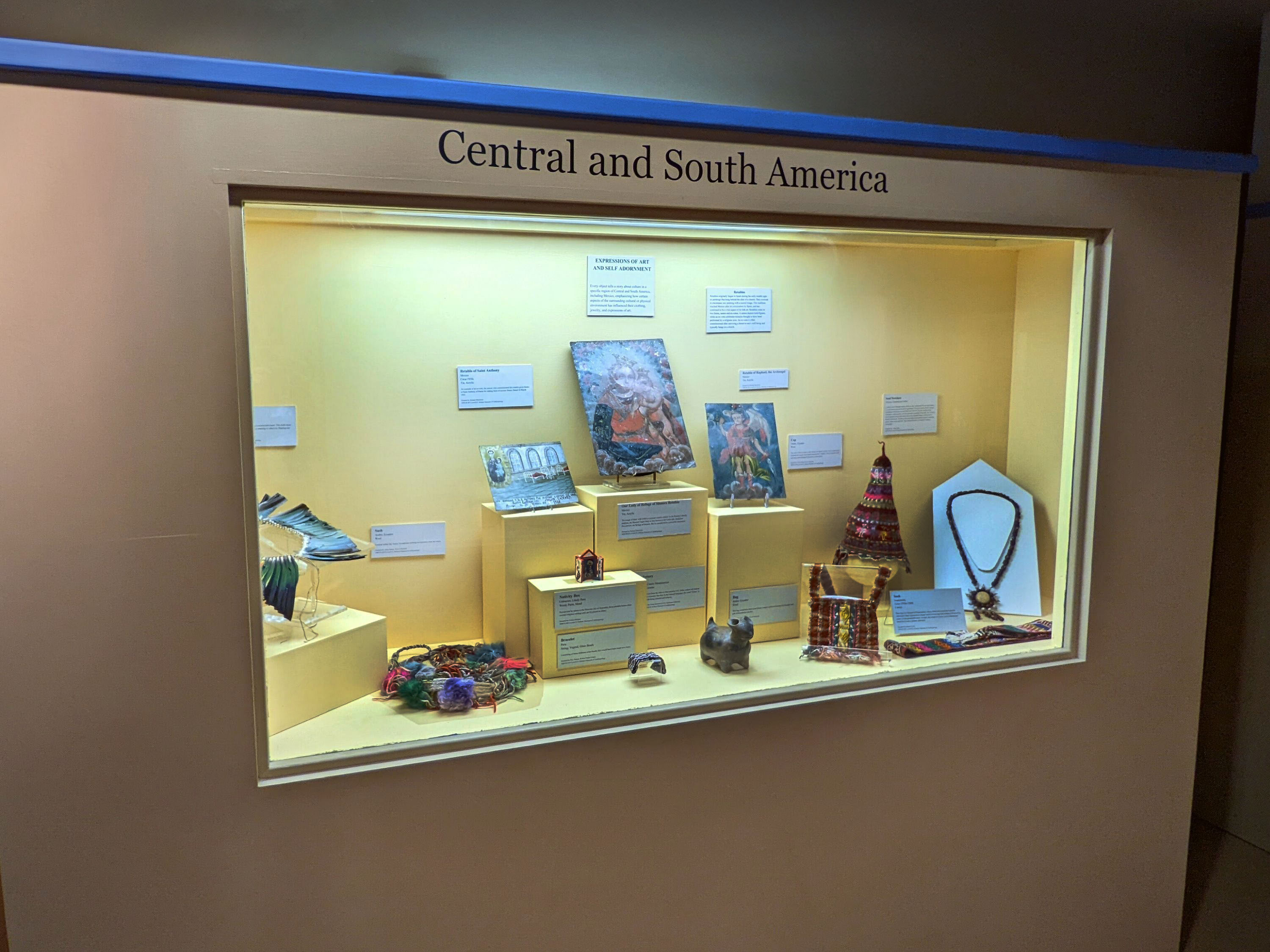 Display case with a yellow background. Objects on display include a feather necklace, sash, metal retablos, pottery of a dog, miniature nativity box, bracelet, and bag. Label attached to walls and on the bottom of the case.