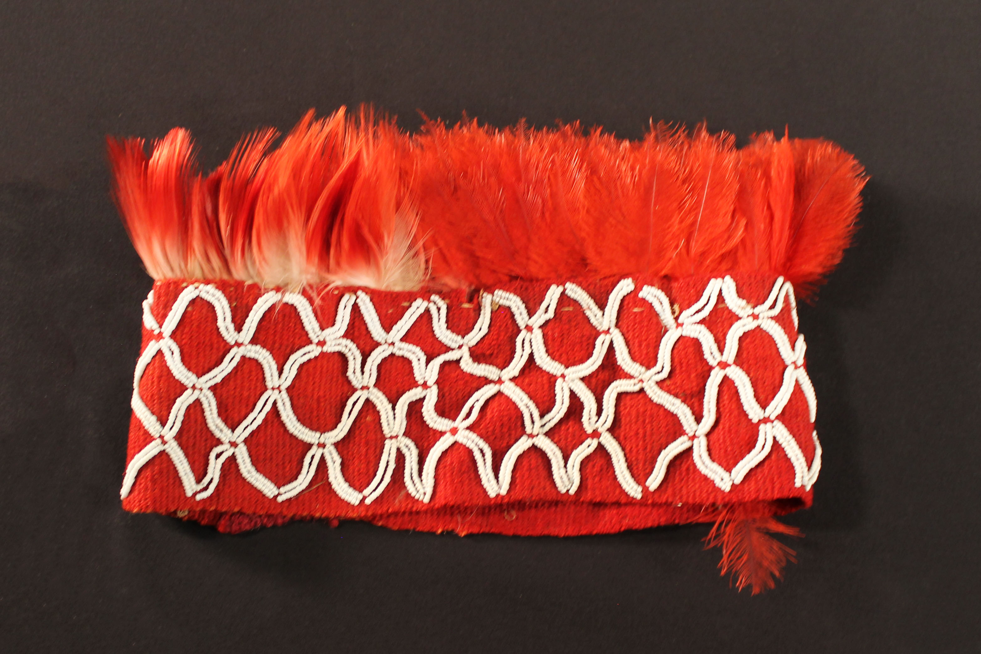 Headdress of red woven material with red feathers sewn to the top of the headband. Body of band decorated with repeated white diamond design. 