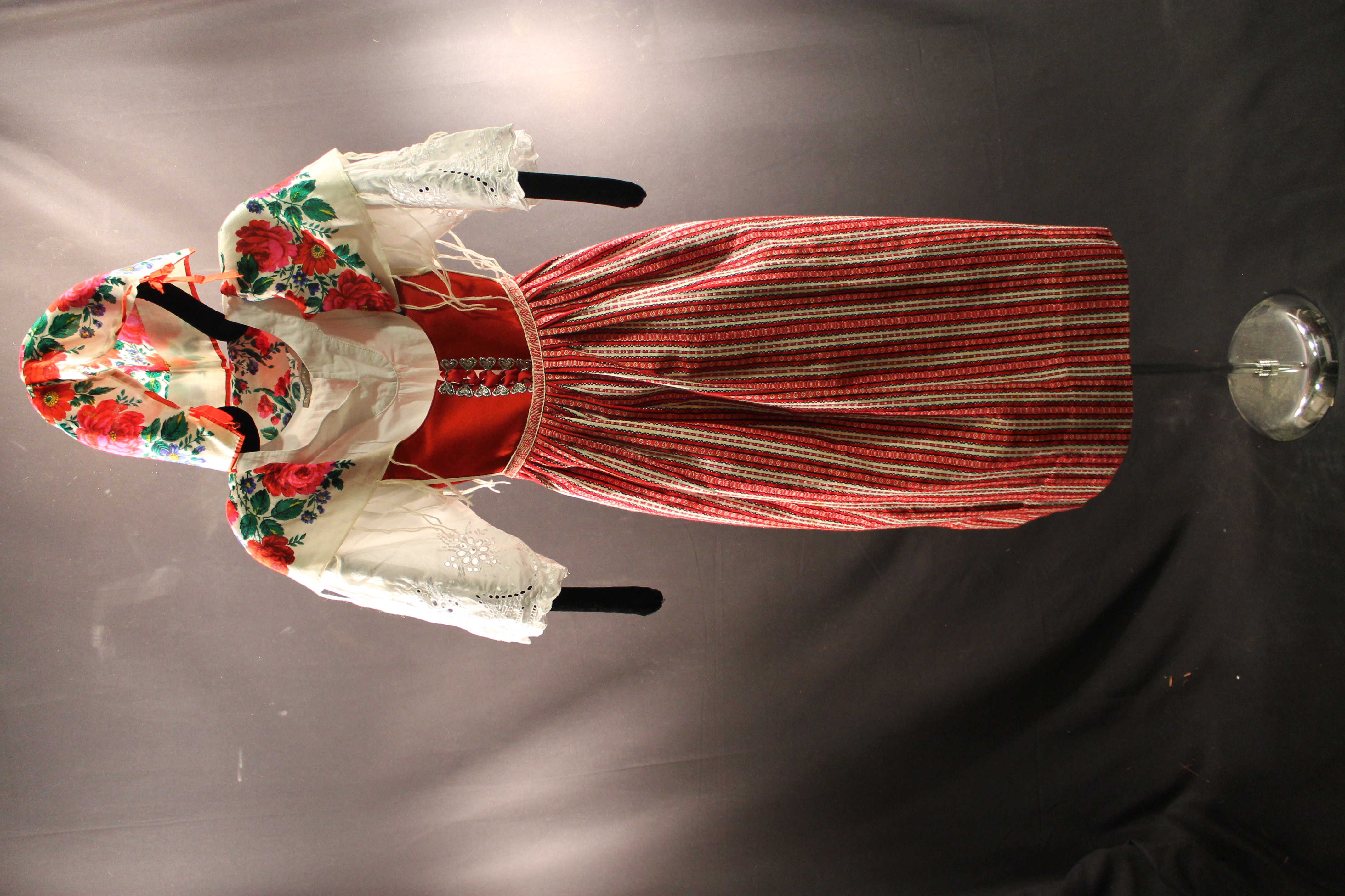 Full Austrian costume with a striped skirt, apron, top, vest, bonnet, and scarf. All follow the same red and white colors. Scarf and bonnet decorated with red roses.