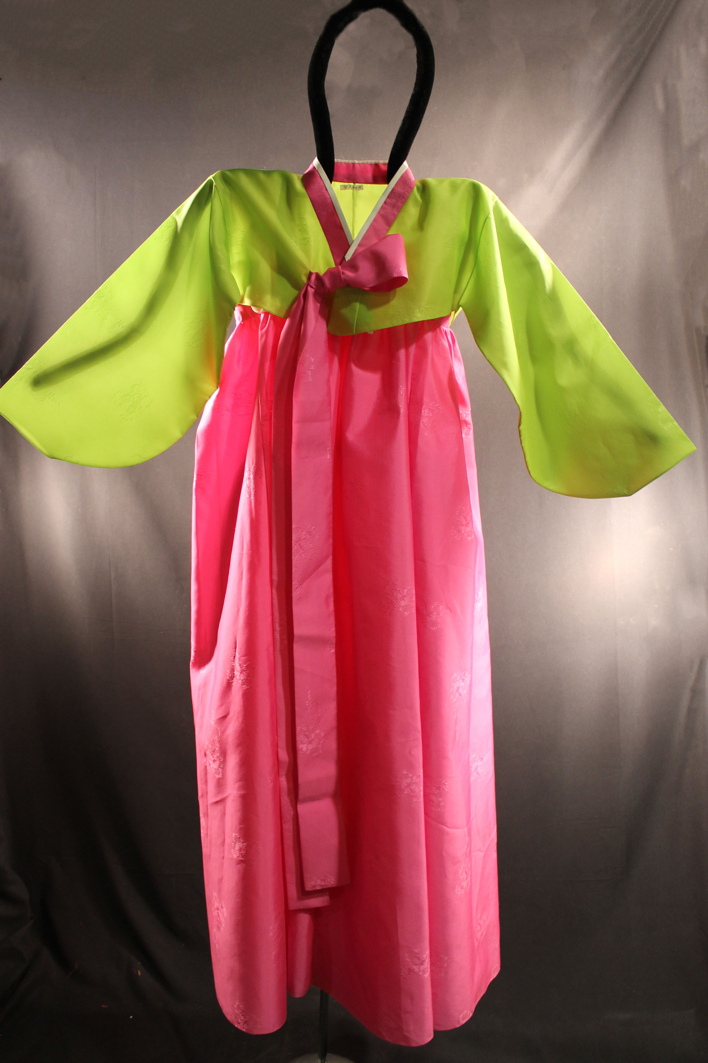 The skirt of the garment is made of hot pink sheer fabric. The jacket is lime green with a hot pink and white belt that is tied into a bow. Both fabrics featured raised floral patterns. 