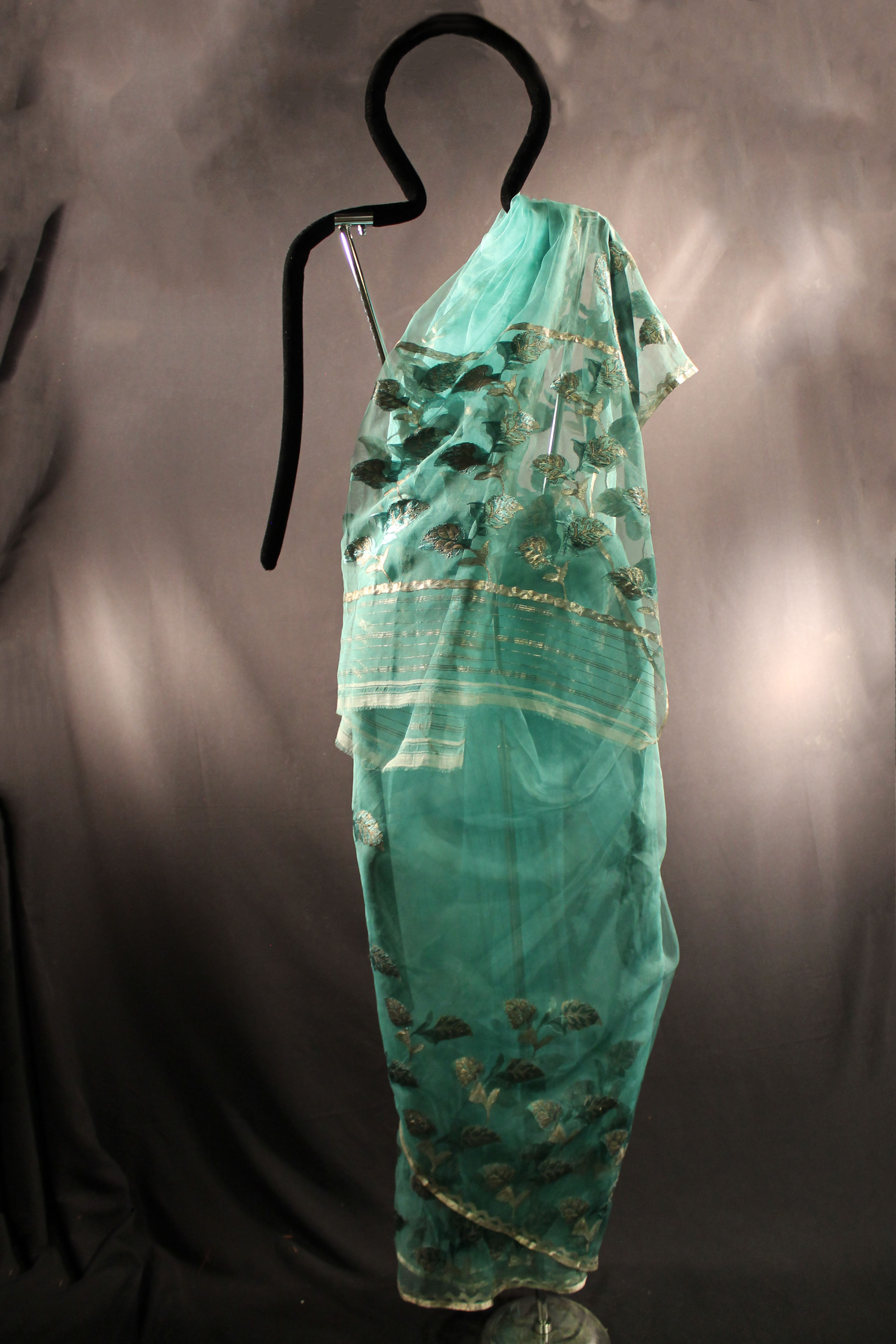 Turquoise cloth sari with edge decorated with silver thread with a zigzag design above it. Also has a leaf pattern design on the body of the garment.