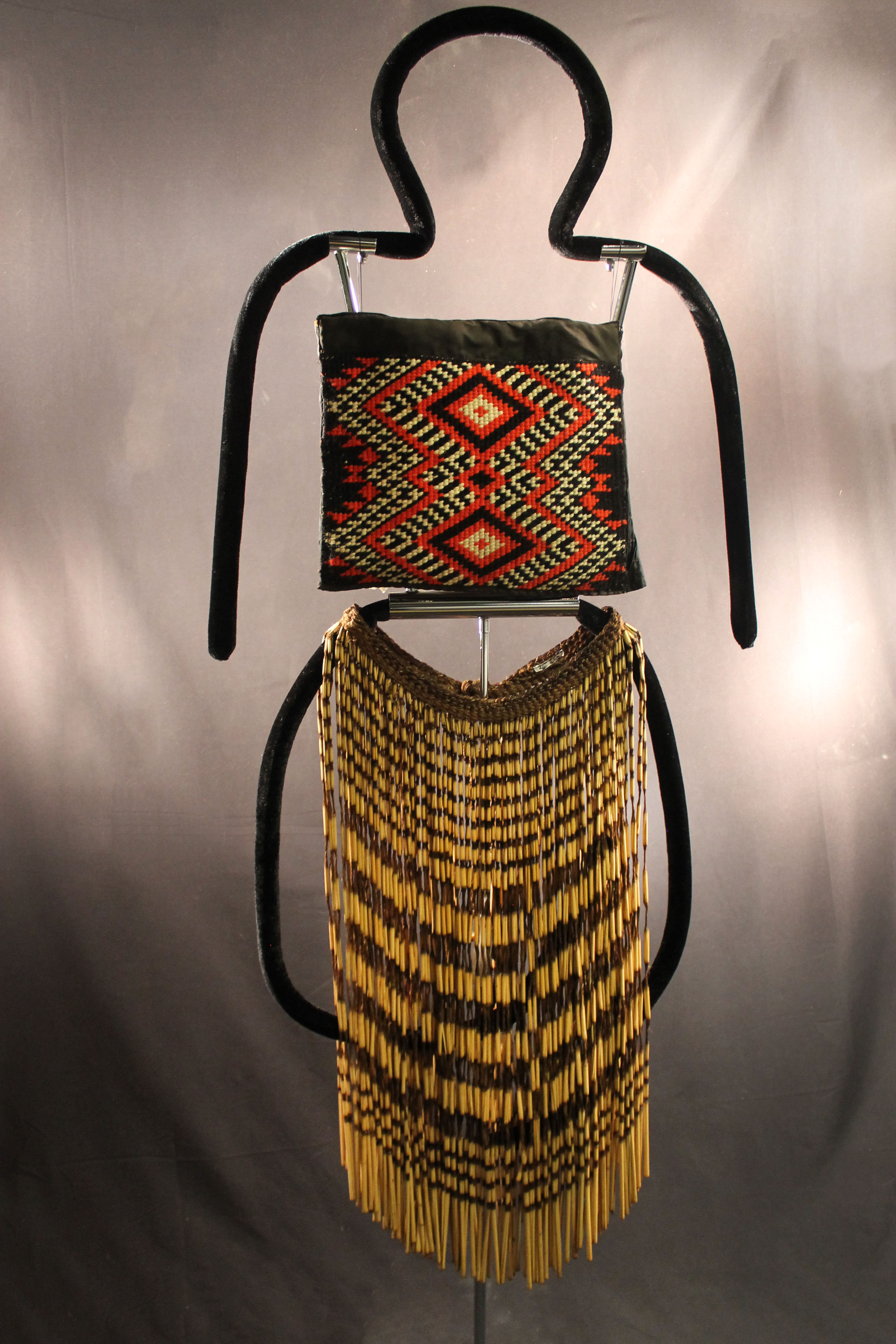 The costume is made up of a top made up of thick fabric with black, white, and red geometric square design. The skirt is tan and brown with reeds held together at the top with braiding.