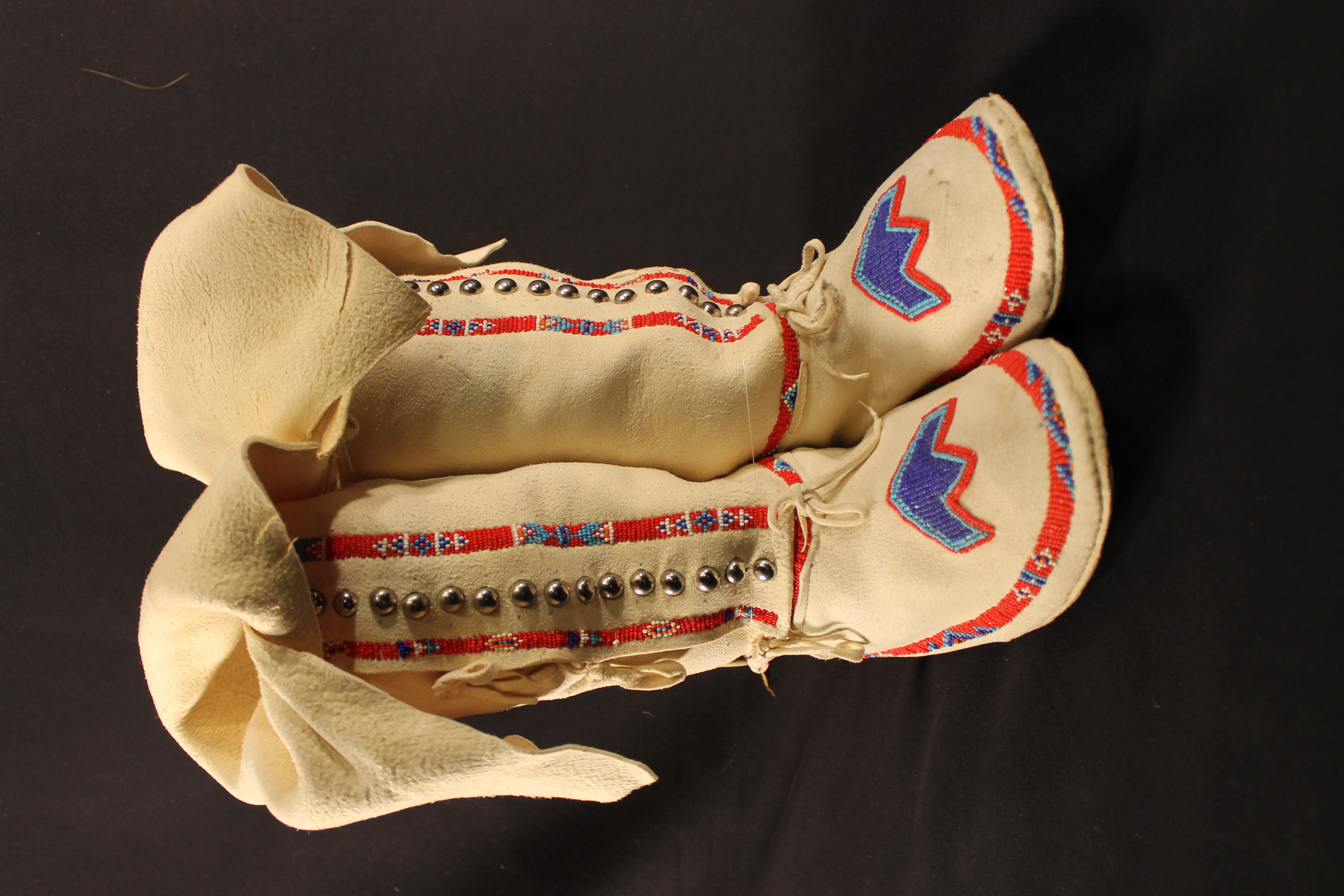 Leather cream-colored moccasin with red, blue, white beadwork. Silver metal studs run down the center. Blue three-pointed geometric beaded design on the toe of the foot.