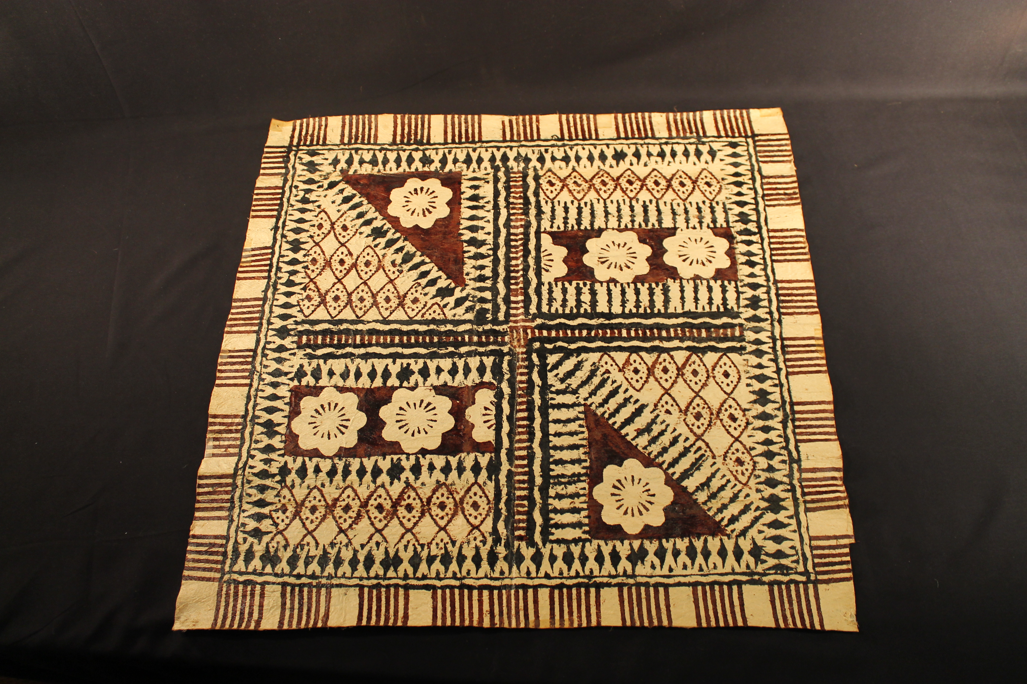 Square bark cloth made of flattened wood with black, red, and maroon-colored designs on a tan background.