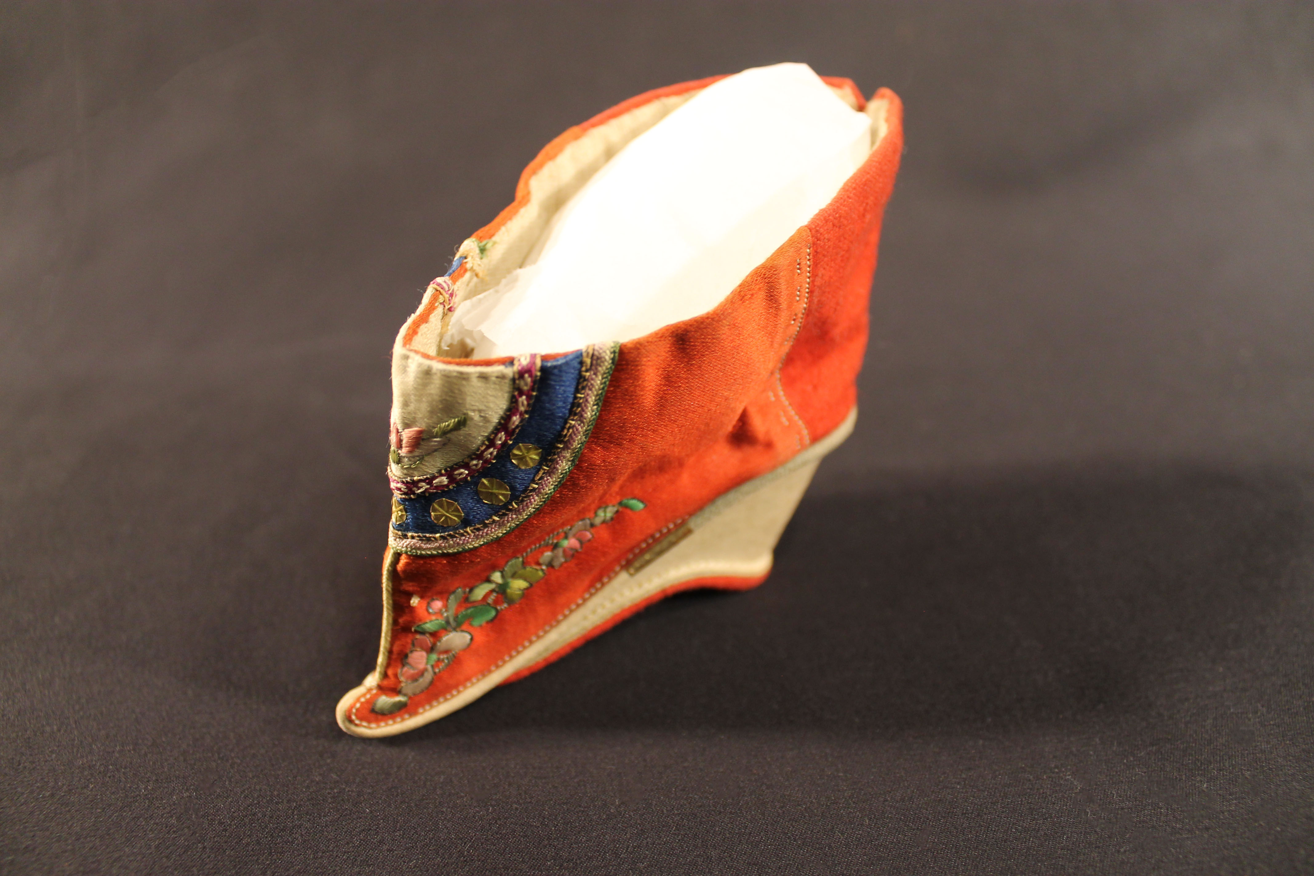 Small shoe with a white base that curves upwards and has a large opening up top. The shoe is primarily red, with flowers and a mandala design on the front.