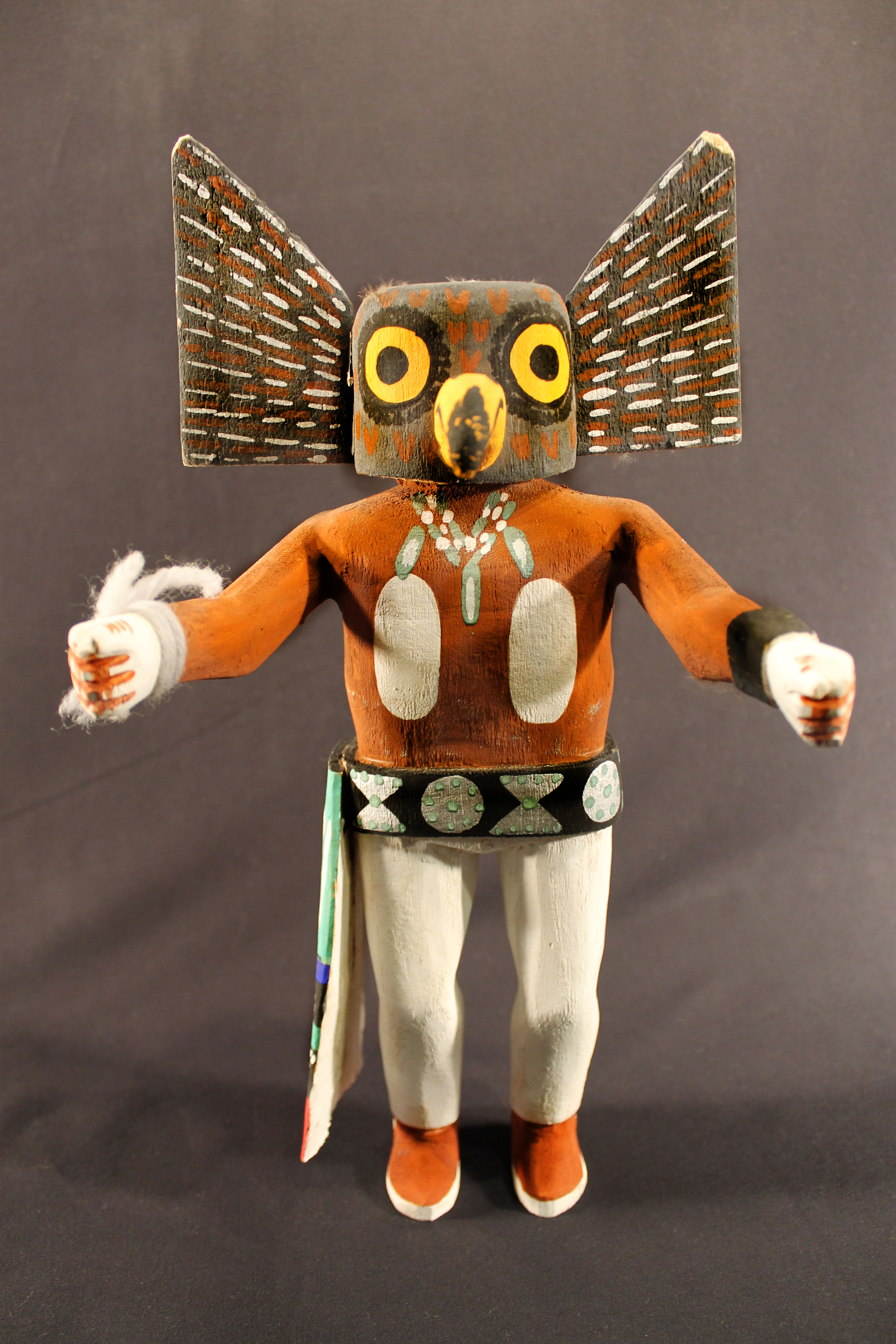 Painted wooden kachina doll that depicts a great horn owl spirit, which has a human body and an owl’s head. It has white-painted pants and a sash that are attached to a silver belt. The doll's upper body is brown with a white shape on the chest and hand and is wearing a painted silver necklace. 